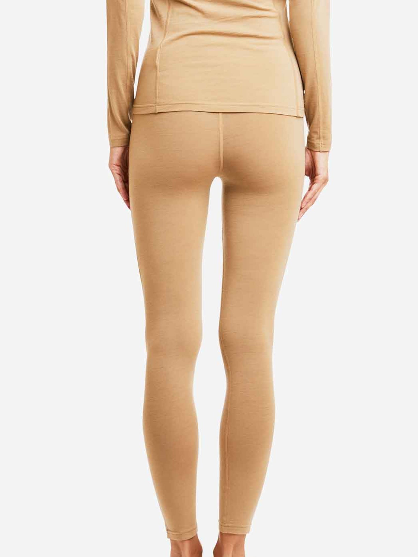 A person is standing with their back to the camera, showcasing the We Norwegians Sno Merino Leggings Women in beige. The outfit includes a matching long-sleeved shirt, both made from super fine merino wool. With their arms relaxed at their sides, the photo emphasizes the lower part of the body against a plain white background.