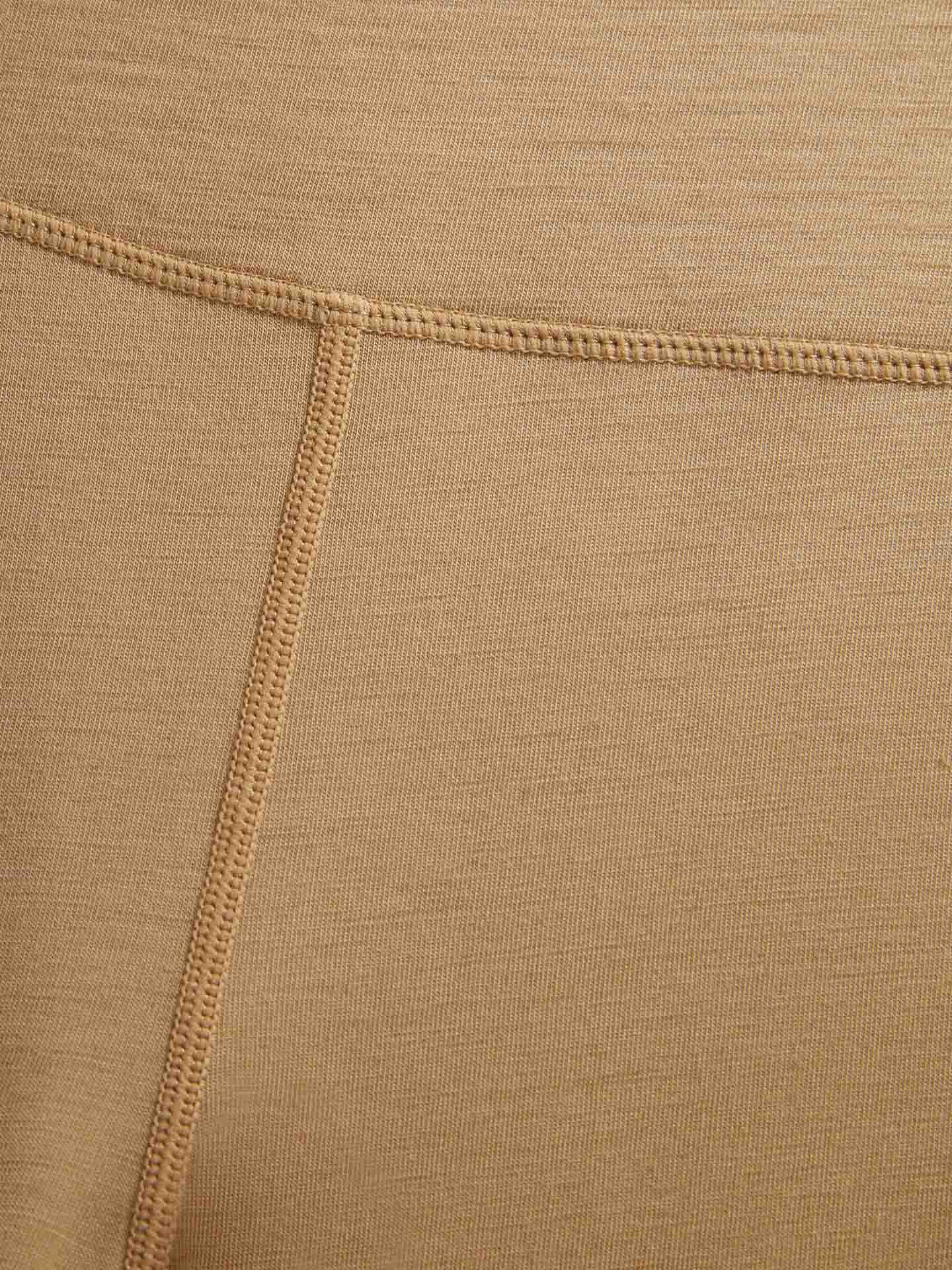 Close-up view of the tan-colored, breathable fabric of the Sno Merino Leggings Women by We Norwegians, showcasing visible stitching. The image focuses on a horizontal seam that intersects with a vertical seam. The texture of the Scandinavian Luxury base-layer fabric and the meticulous weave of the stitches are clearly evident.