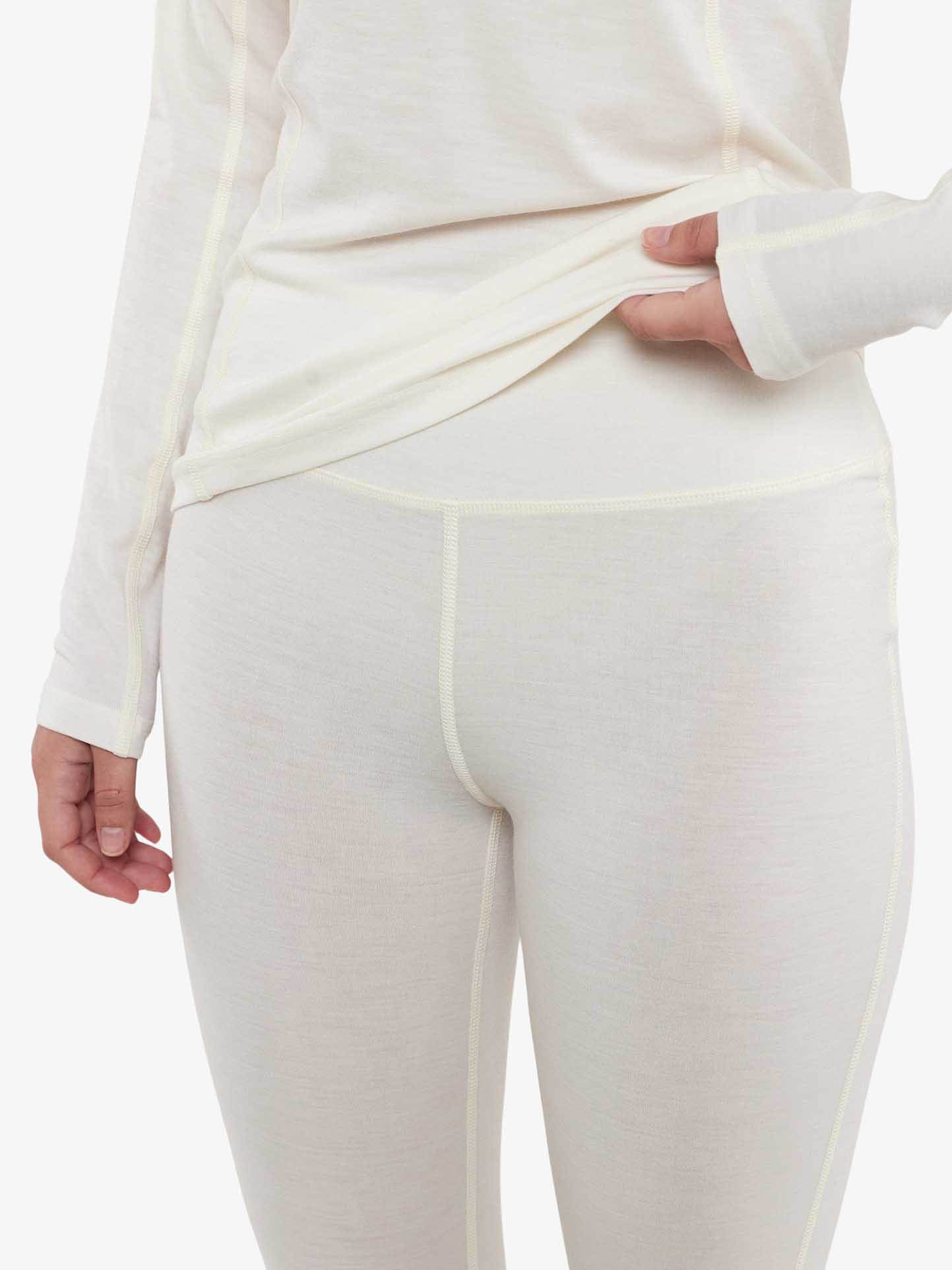 A woman is photographed from the back, wearing a beige long-sleeve top and Sno Merino Leggings by We Norwegians, and gently lifting the hem of her top to reveal the leggings' waistband. The outfit, crafted from super fine merino wool, highlights its soft, comfortable, and breathable fabric.