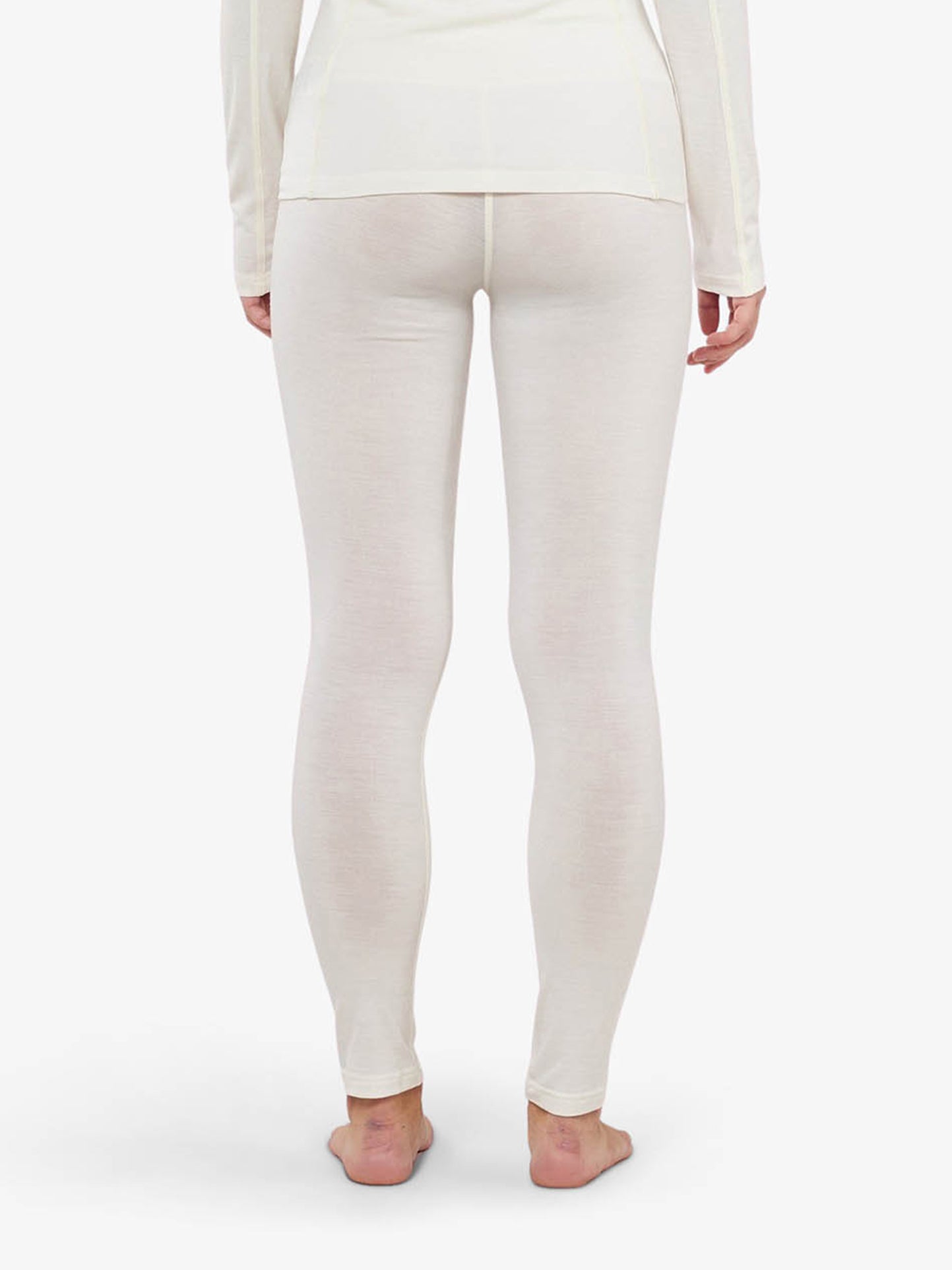 A woman dressed in an off-white, fitted long-sleeve top and matching Sno Merino Leggings from We Norwegians is shown from the back, standing barefoot against a plain white background. These Scandinavian luxury base-layers appear to be made of a light, breathable fabric suitable for layering or lounging.