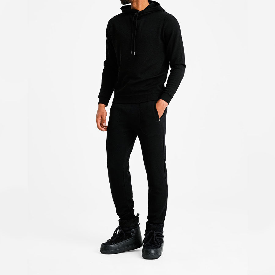 A person is standing and wearing a Tind Hoodie Men Black from We Norwegians, crafted from breathable and antibacterial Italian Yarn. They are also dressed in matching black jogger pants and black high-top sneakers. The person has their hands in their pockets against a plain, white background, and their face is not visible.