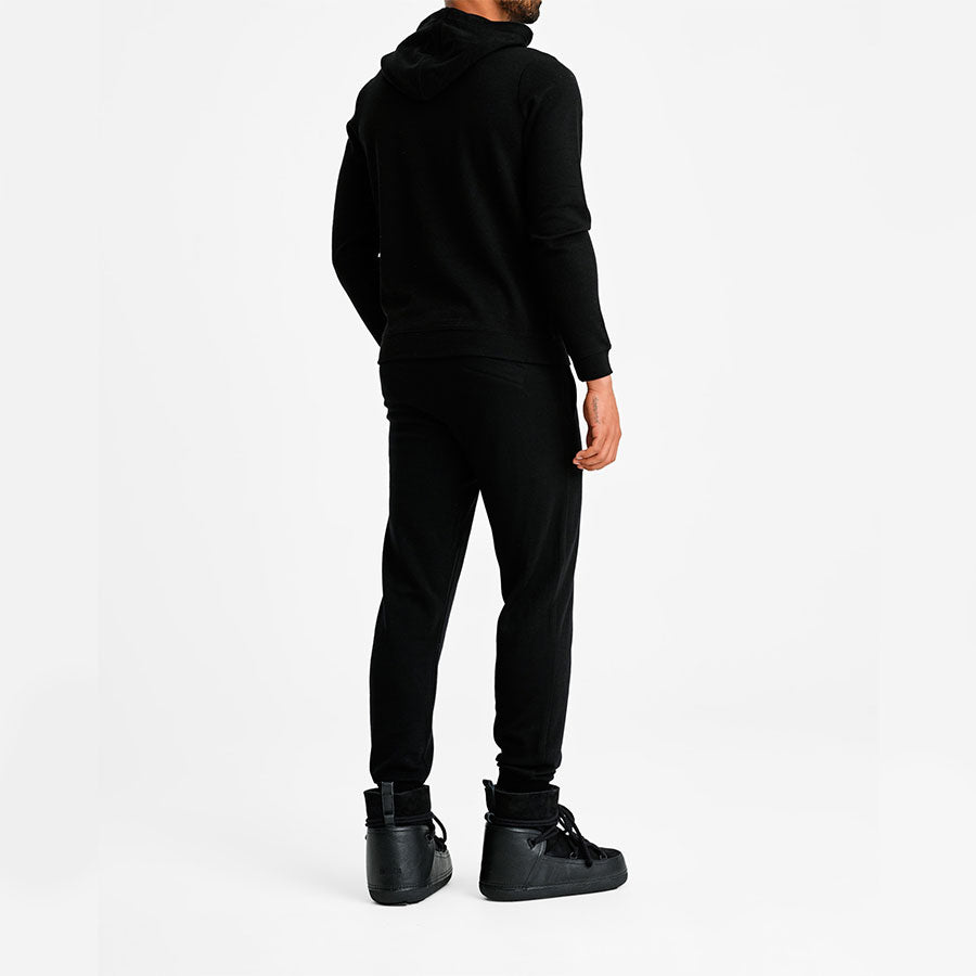 A person dressed in a Tind Hoodie Men Black by We Norwegians, black pants, and black sneakers stands facing away from the camera against a plain white background. The outfit is casual, monochromatic, and crafted for elegance with breathable and antibacterial properties.