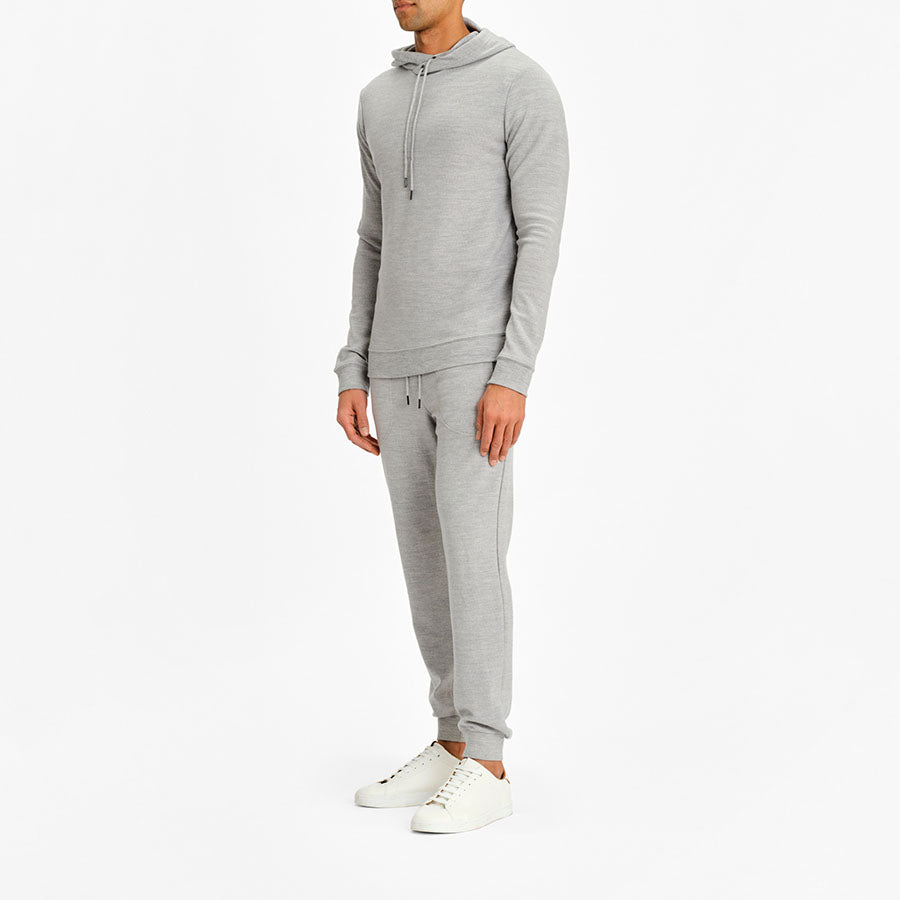 A person is modeling the Limited Time Bundle: Tind Hoodie & Tind Joggers Men by We Norwegians, a light gray set perfect for any casual wardrobe. The outfit is complemented by clean, white sneakers, and they are standing against a plain white background.