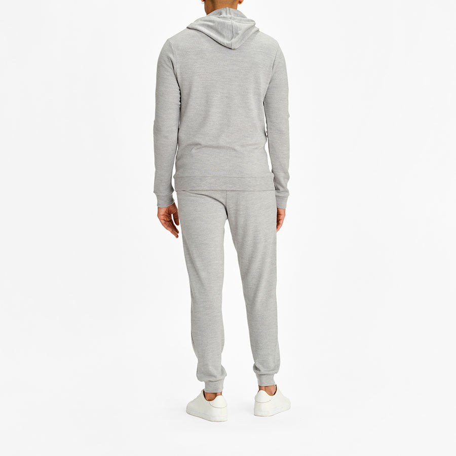 A person wearing the We Norwegians Tind Hoodie Men and matching grey sweatpants, both crafted from Italian Spun Superfine Merino, stands facing away from the camera. The outfit is complemented by white sneakers. The background is plain white.