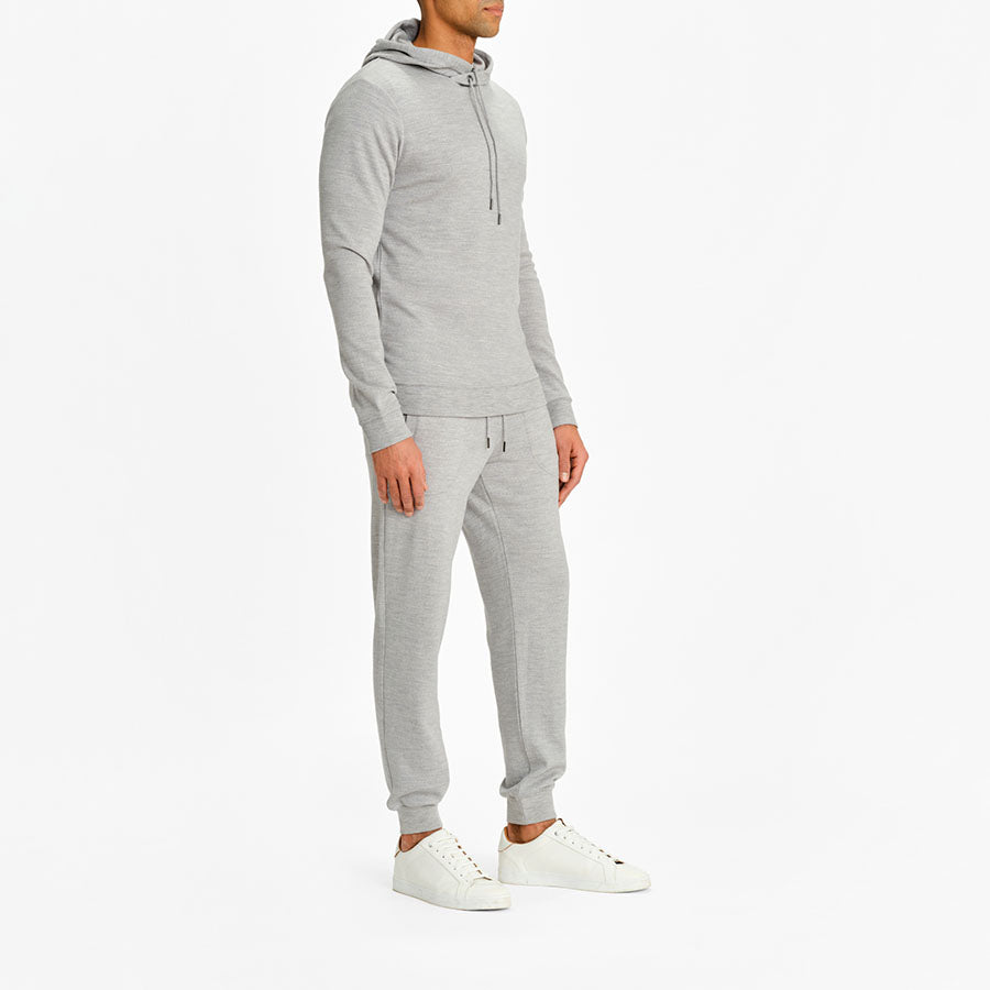 A person is standing against a plain white background, wearing a matching gray Tind Hoodie Men from We Norwegians and sweatpants. The hoodie, made of Superfine Merino wool, has a drawstring, and the sweatpants feature pockets and a drawstring waist. The person completes the look with white sneakers.