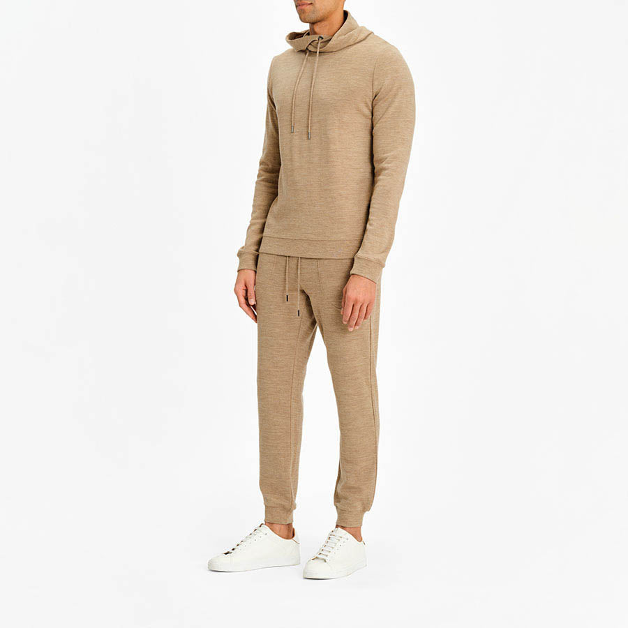 A person stands against a plain white background, dressed in the Tind Hoodie Men Camel from We Norwegians along with matching jogger pants featuring drawstrings. The outfit is crafted from breathable and antibacterial Italian Yarn. They are wearing white sneakers. The person's face is not visible.