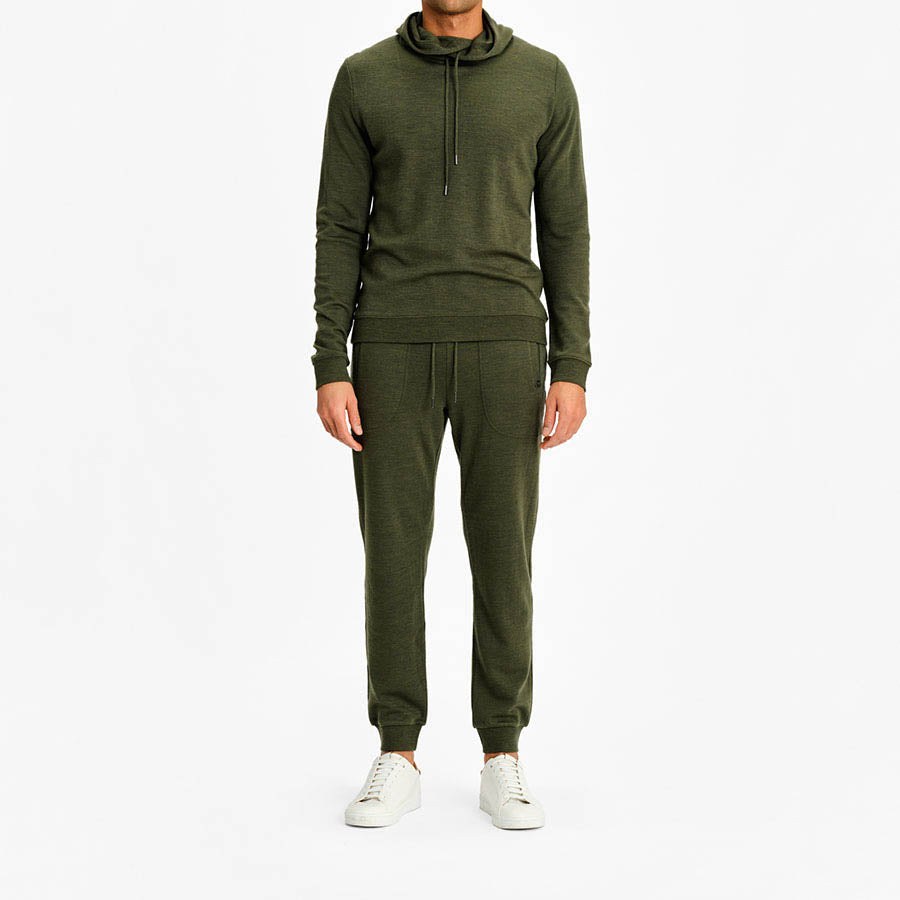 A person is standing against a plain white background, wearing an olive green Tind Hoodie Men from We Norwegians, made from superfine merino wool, along with matching sweatpants. Their white sneakers complement the relaxed position of their hands at their sides. The outfit exudes comfort and casual style.