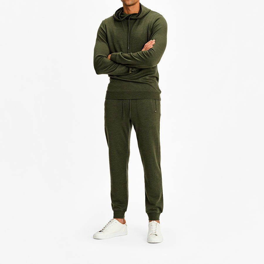 A person stands against a white background, arms crossed. They wear an olive green Tind Hoodie Men from We Norwegians, made from superfine merino, along with matching jogger pants and white sneakers. The head is out of the frame.