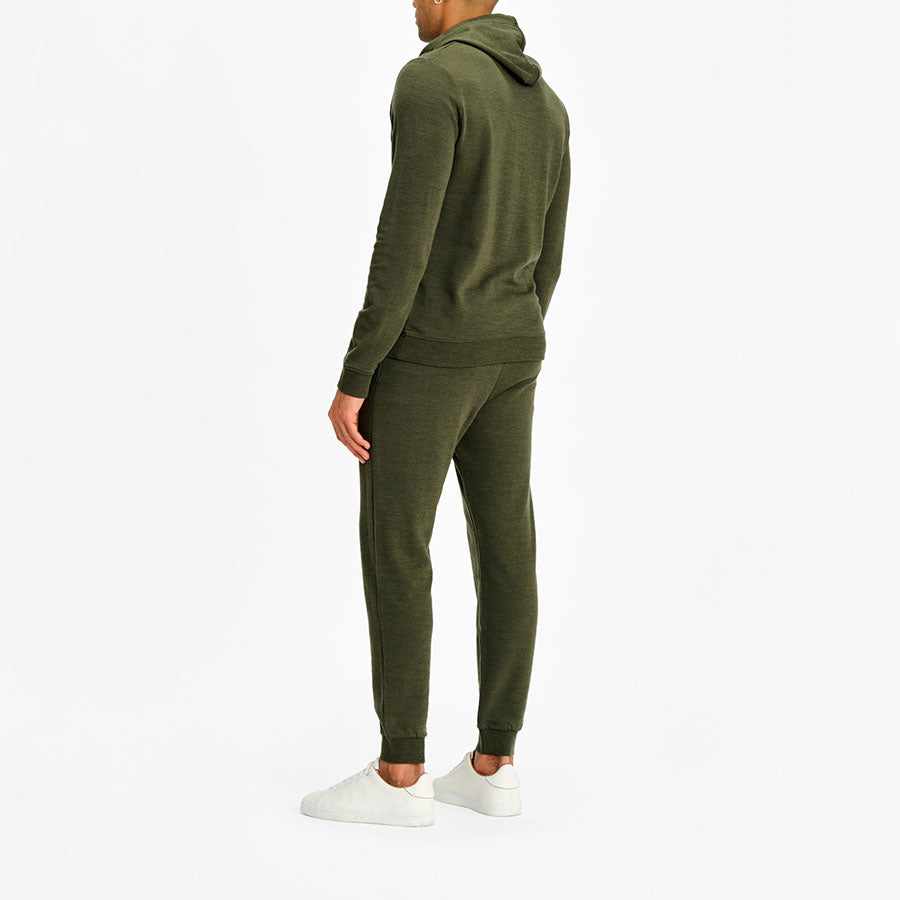 A person is shown from the back, dressed in a matching dark green Tind Hoodie Men Olive Green by We Norwegians and jogger pants made from superfine Merino wool. They are standing upright, with one arm relaxed by their side and the other partially visible. They are wearing white sneakers, and the background is plain white.