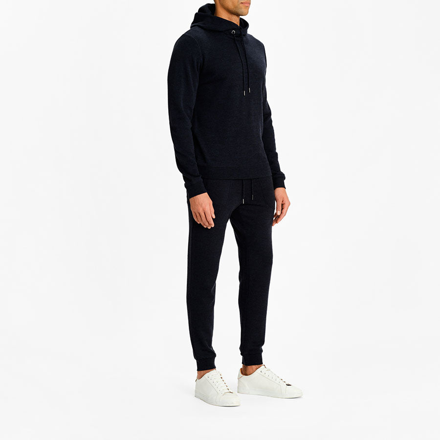 A person in We Norwegians' black Tind Hoodie Men, crafted from superfine Merino, and matching black joggers stands against a white background. The individual, also wearing white sneakers, has their face hidden. The hoodie features drawstrings, and the joggers have elastic cuffs at the ankles.