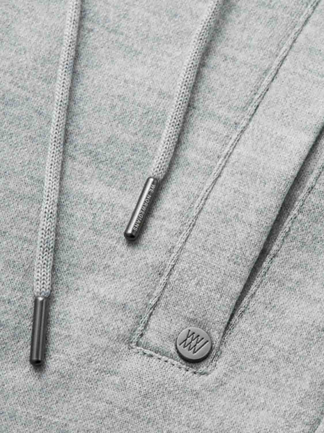 Close-up of a grey hoodie showing the drawstring ends, a part of the pocket, and a small metal button engraved with a logo. The fabric appears to be soft and thick, suggesting warmth and comfort, reminiscent of the contemporary-cool designs found in We Norwegians' Tind Joggers for Men.