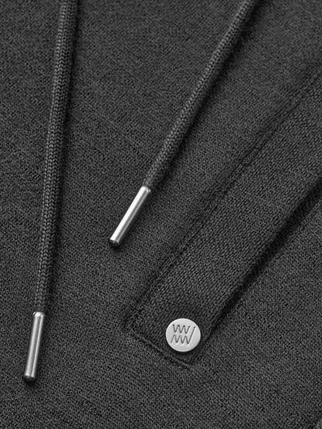 Close-up of the details on a dark gray fabric piece, potentially from the Tind Joggers Men by We Norwegians. It features two drawstrings with metal tips and a small round metallic logo tag engraved with "WW." The texture of the fabric and stitching are visible, reflecting contemporary-cool designs typical of Merino wool joggers.