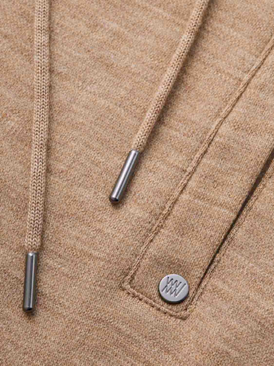 Close-up of a beige hoodie showing the drawstrings and a small metallic button attached near the pocket. The drawstrings are tipped with metallic caps, and the button features an engraved design. The fabric appears soft and textured, perfectly complementing We Norwegians' Tind Joggers Men Camel for contemporary-cool designs.