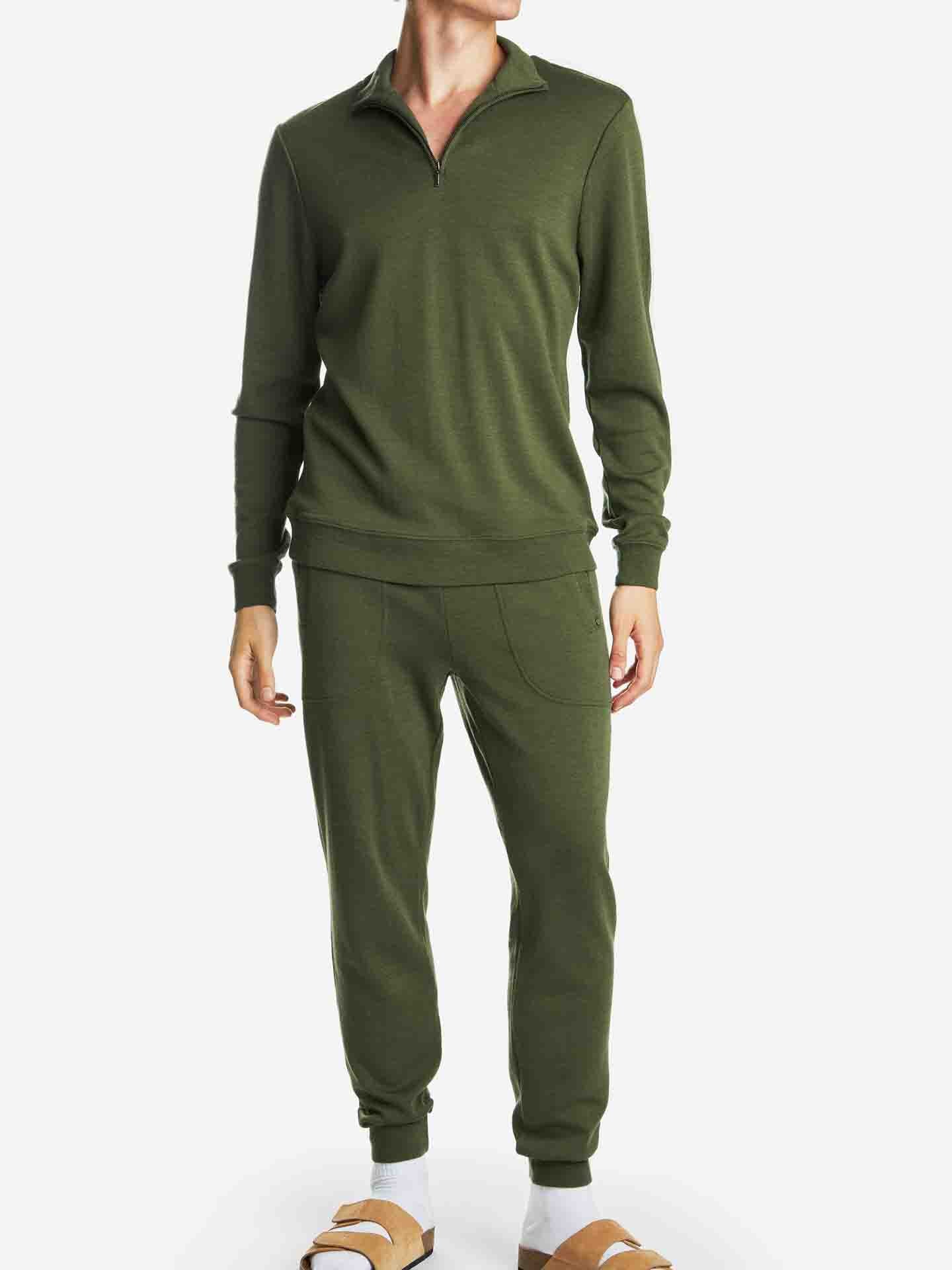 A person is wearing the Tind Joggers Men in Olive Green by We Norwegians, featuring a long-sleeve, half-zip top and matching relaxed fit jogger-style pants. The outfit is paired with light brown sandals. The background is white, and the person's face is not visible.