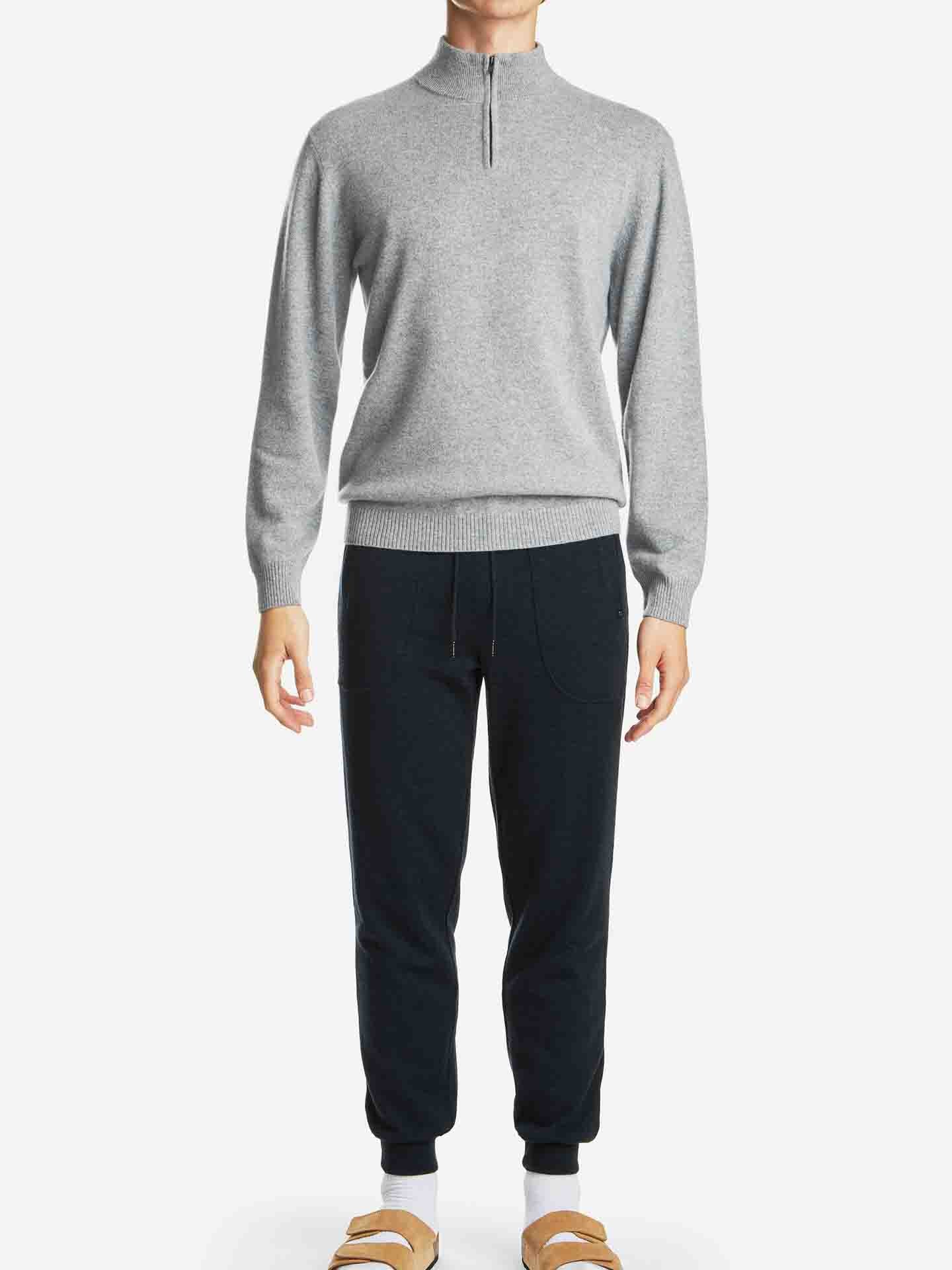 A person is wearing a light grey quarter-zip sweater with long sleeves and dark blue We Norwegians Tind Joggers Men, which are antibacterial and odor-resistant. They are also wearing light brown sandals. The person's face is not visible in the image, and the background is solid white.