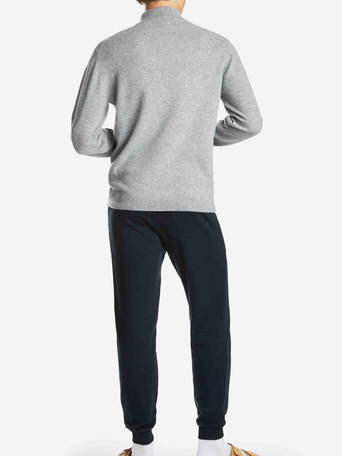 A person stands facing away from the camera, wearing a light grey sweater and dark navy Tind Joggers Men by We Norwegians. Their attire, featuring contemporary-cool designs, is completed with white socks and tan shoes against a clean white background.