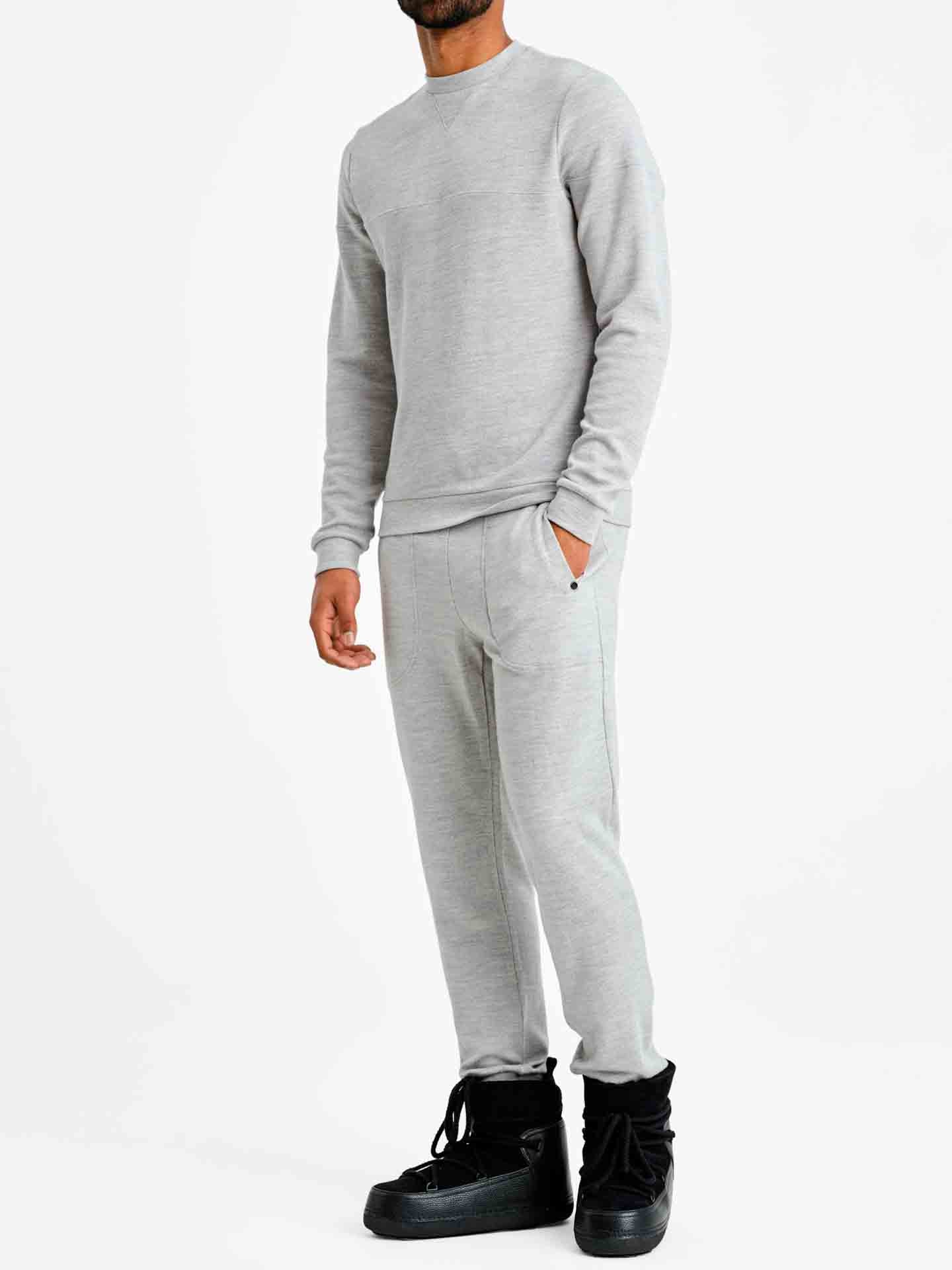 A person wearing the Limited Time Bundle: Tind Crewneck & Tind Joggers Men by We Norwegians, featuring a light gray, long-sleeve sweatshirt and matching light gray Merino wool joggers, stands against a white background. They are also wearing black high-top sneakers. The person's face is not visible in the image.