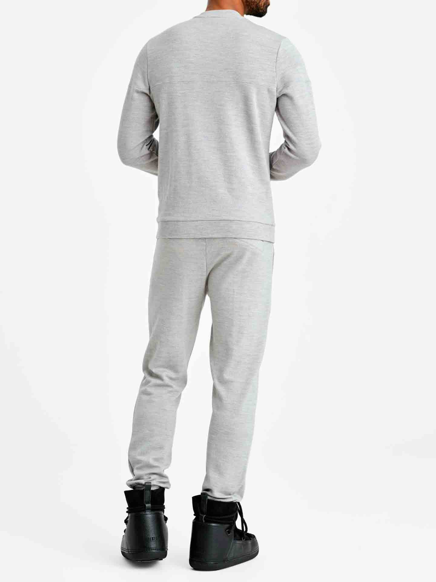 A person is standing facing away from the camera wearing a light gray We Norwegians tracksuit paired with black sneakers. The background is plain white. The tracksuit, which includes a long-sleeved top and matching Tind Joggers Men pants, showcases contemporary-cool designs.