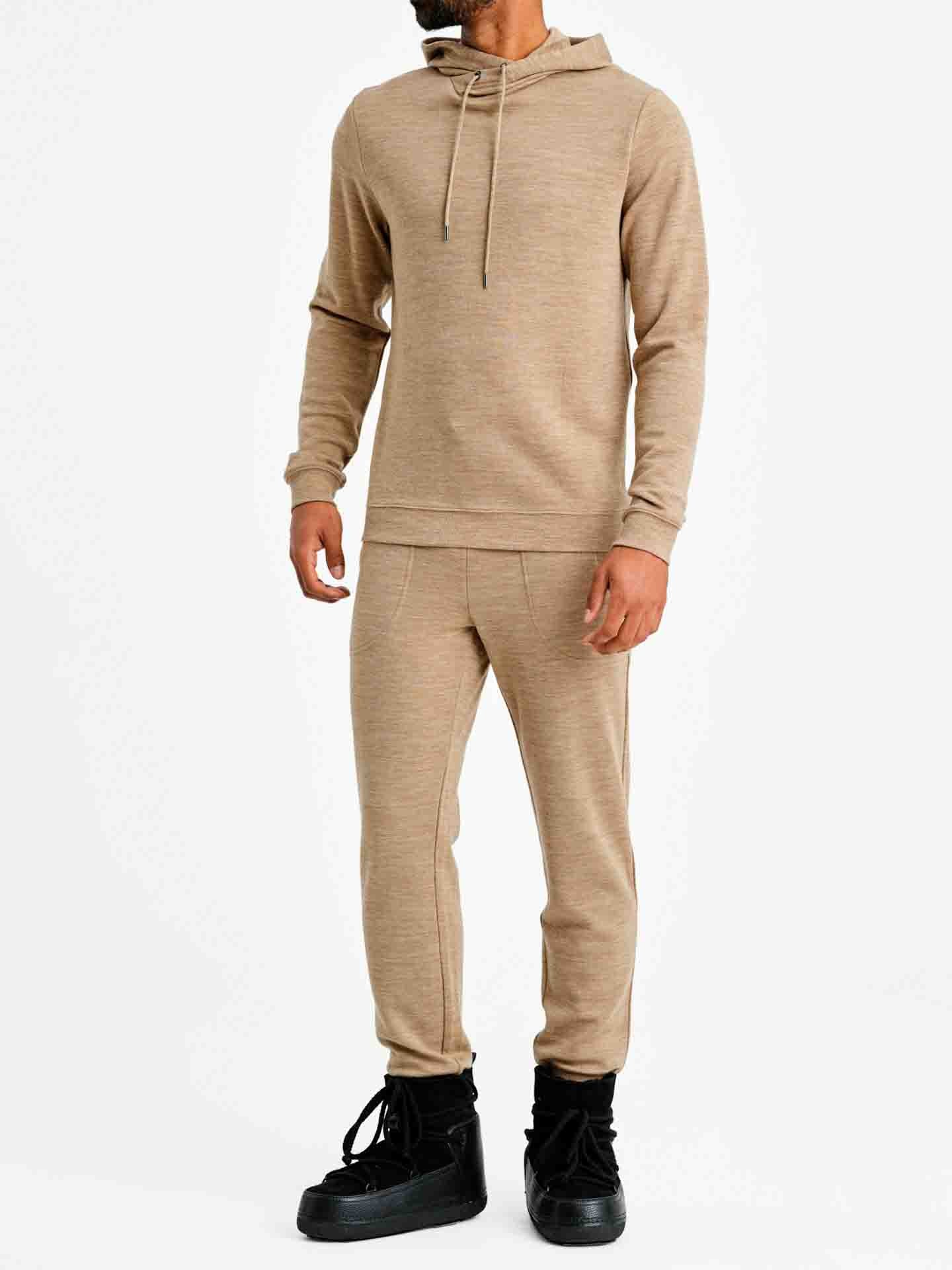 A person is standing against a white background, wearing a beige hoodie with drawstrings and contemporary-cool, slouchy Tind Joggers Men Camel by We Norwegians in matching beige. They are also sporting black high-top sneakers. The person's hands are relaxed by their sides.