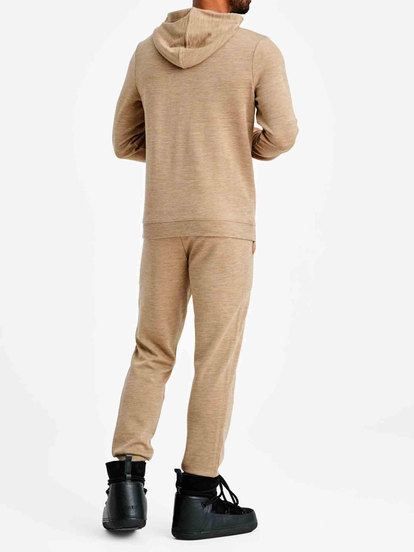 A person is standing against a white background, wearing the Tind Joggers Men Camel by We Norwegians along with a matching beige hoodie. The person is facing away from the camera, highlighting the contemporary-cool design of the outfit. Black boots complement this casual attire.