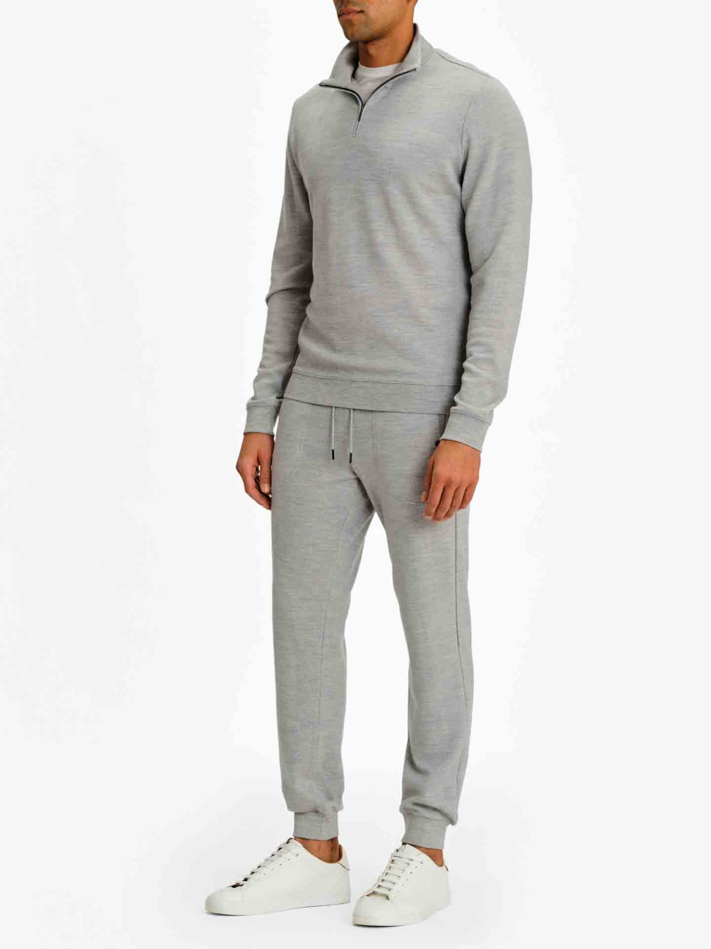 A person stands against a plain white background wearing the Tind 1/4 Zip Sweater Men Grey Melange by We Norwegians, a light grey, long-sleeved pullover sweatshirt made from superfine Merino, paired with matching joggers. They also have on white sneakers. The clothing appears casual and comfortable. The face of the person is not visible in the image.