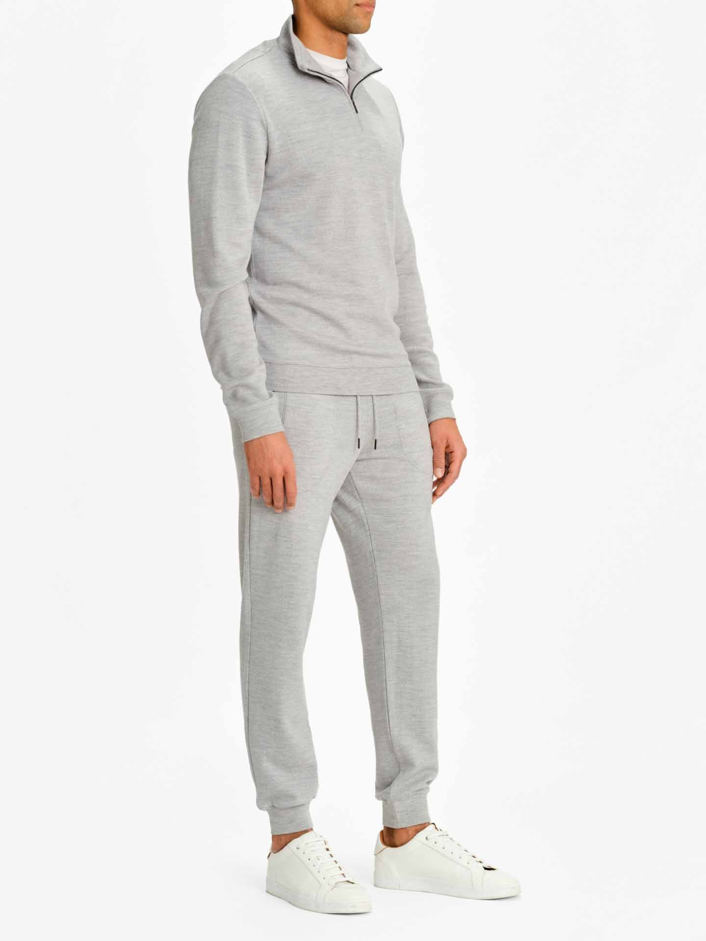 A person stands wearing a light gray Tind 1/4 Zip Sweater Men Grey Melange from We Norwegians, paired with matching joggers and white sneakers. The individual is turned slightly to the side, revealing the front and side profile of the outfit against a plain white background.