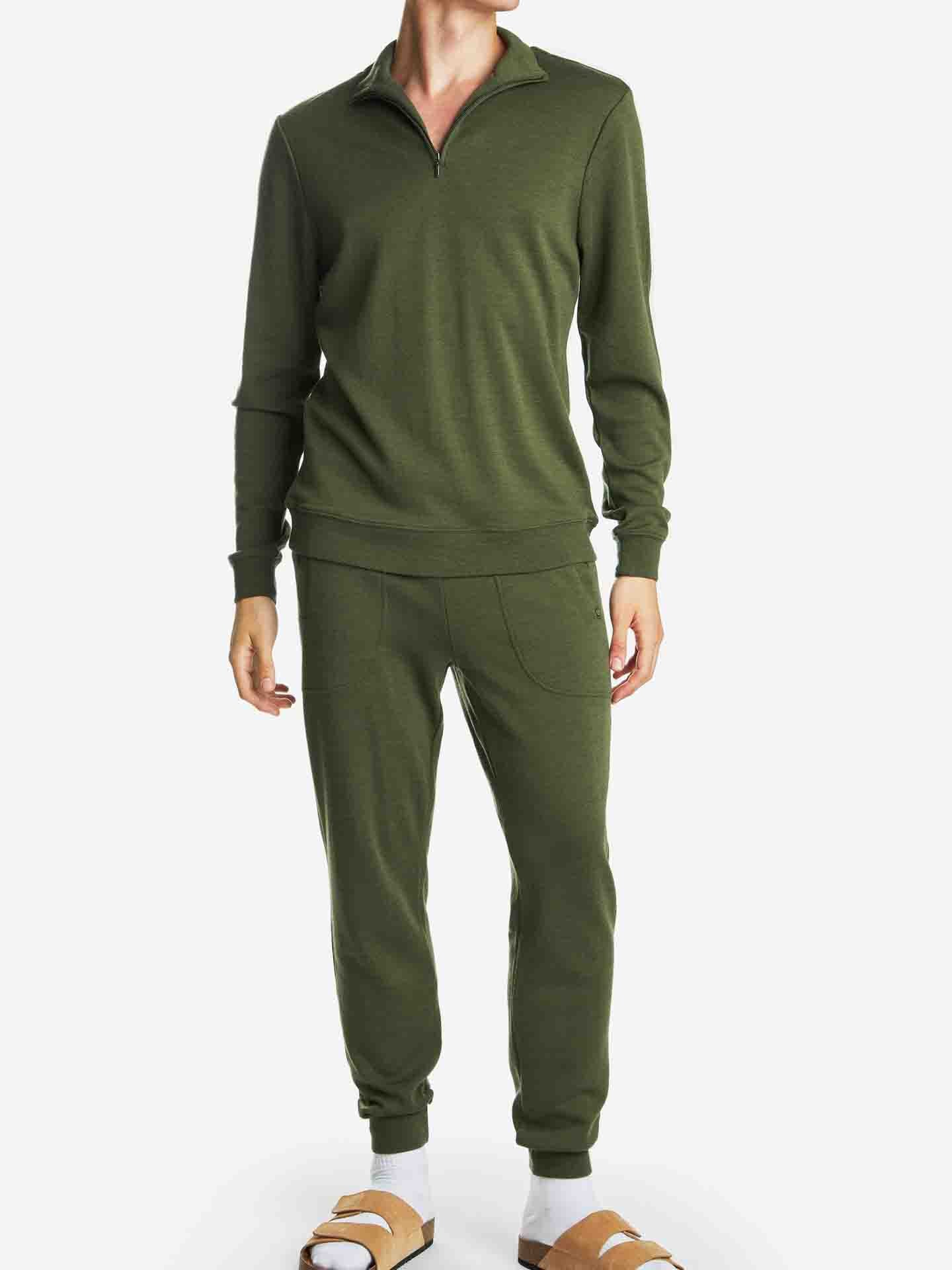 A person is wearing a matching olive green outfit consisting of the Tind 1/4 Zip Sweater Men Olive Green by We Norwegians and jogger pants. Their feet are in tan-colored sandals. They are standing against a plain white background.