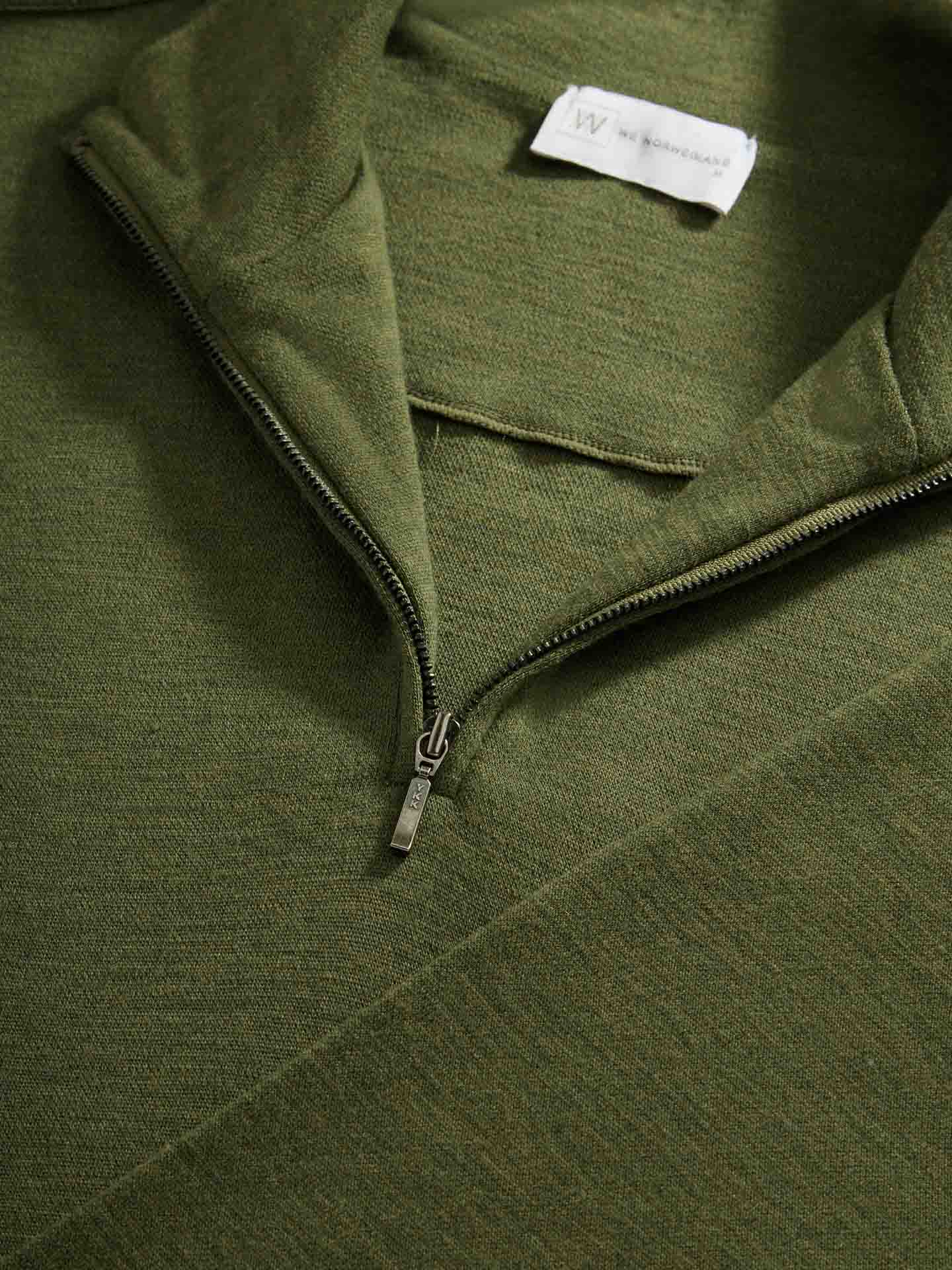 A close-up of the Olive Green Tind 1/4 Zip Sweater for men by We Norwegians lying flat. The partially unzipped collar, featuring a YKK zipper, reveals a tag inside the garment. Although the text and small logo on the tag are not fully readable in the image, the slightly textured weave of the fabric is clearly visible.