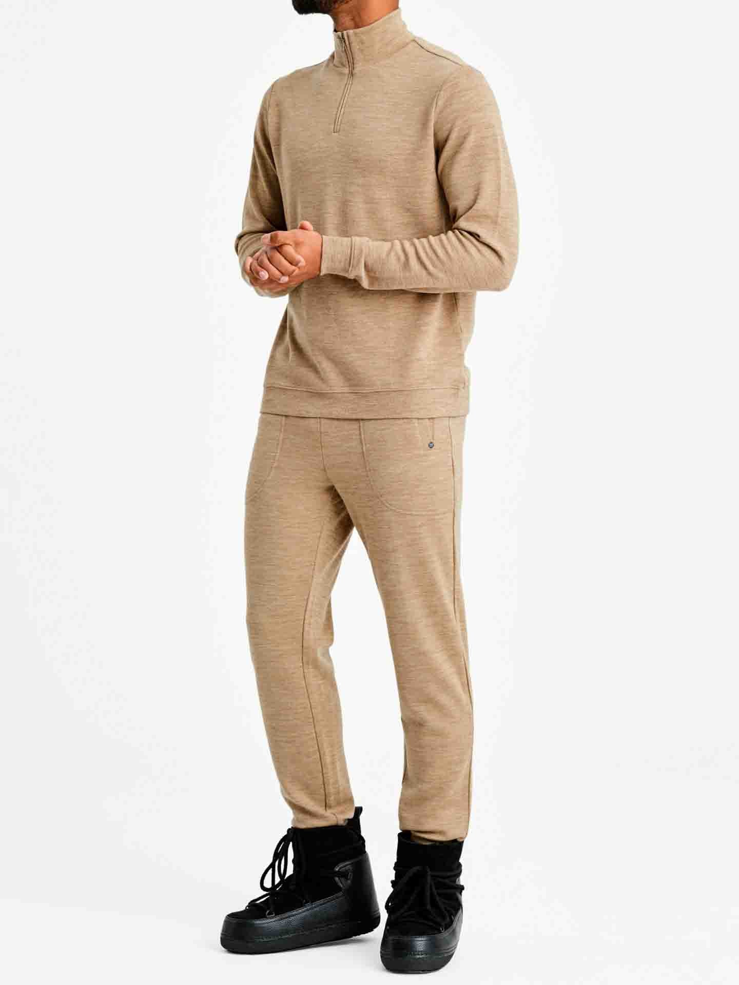 A person stands against a plain white background, wearing a matching beige jacquard knit garment—a long-sleeve quarter-zip top (Tind 1/4 Zip Sweater Men Camel by We Norwegians) paired with Tind Pant matching trims. The person has their hands clasped and is wearing black high-top shoes.