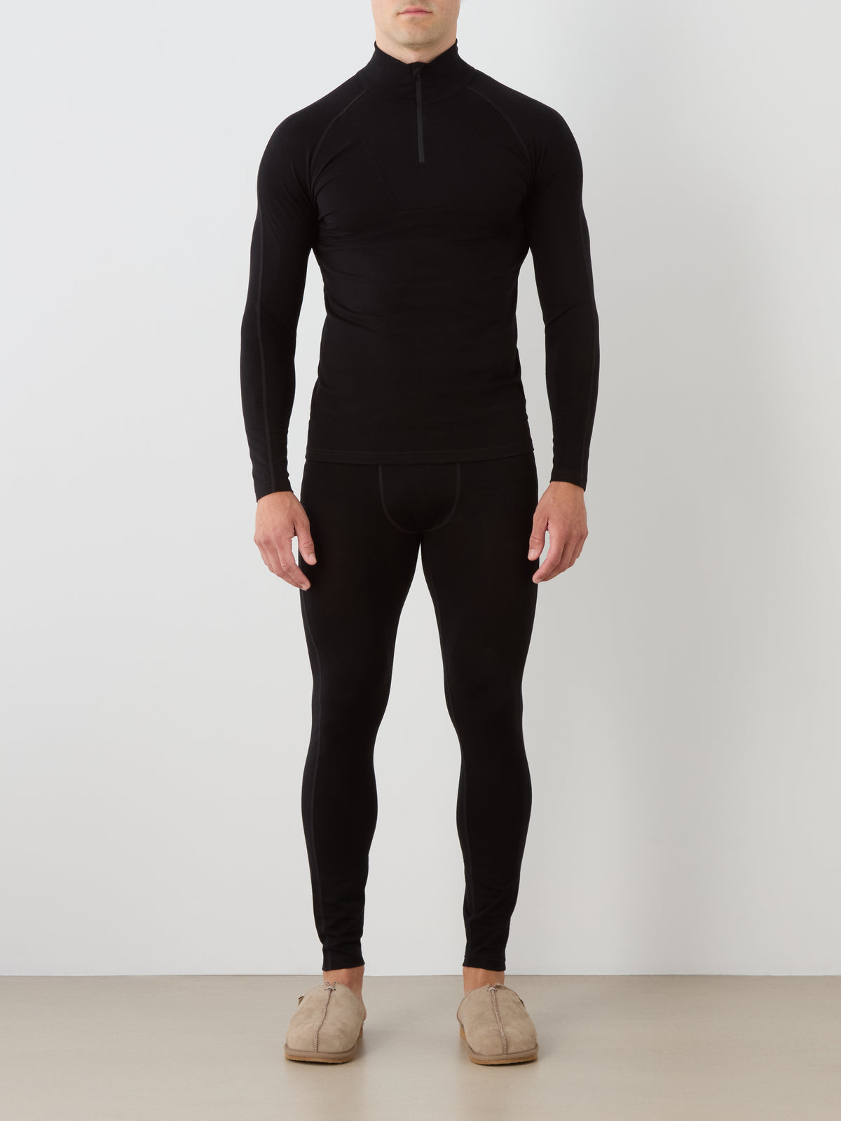 A person stands against a neutral background wearing the We Norwegians Sno Merino Zip Up Men top in black, paired with matching black leggings. They are also wearing tan, slip-on suede slippers.