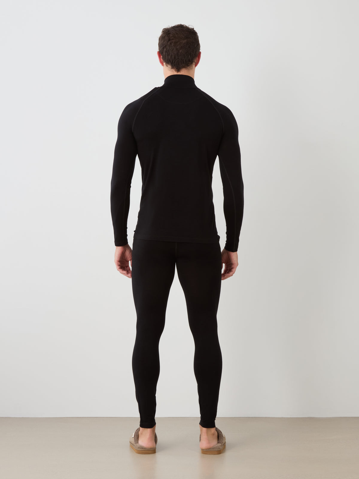 A man stands facing away from the camera, wearing the We Norwegians Sno Merino Zip Up Men base layer top and matching leggings in black, paired with brown slippers, in a minimal indoor setting with a light floor and white wall.
