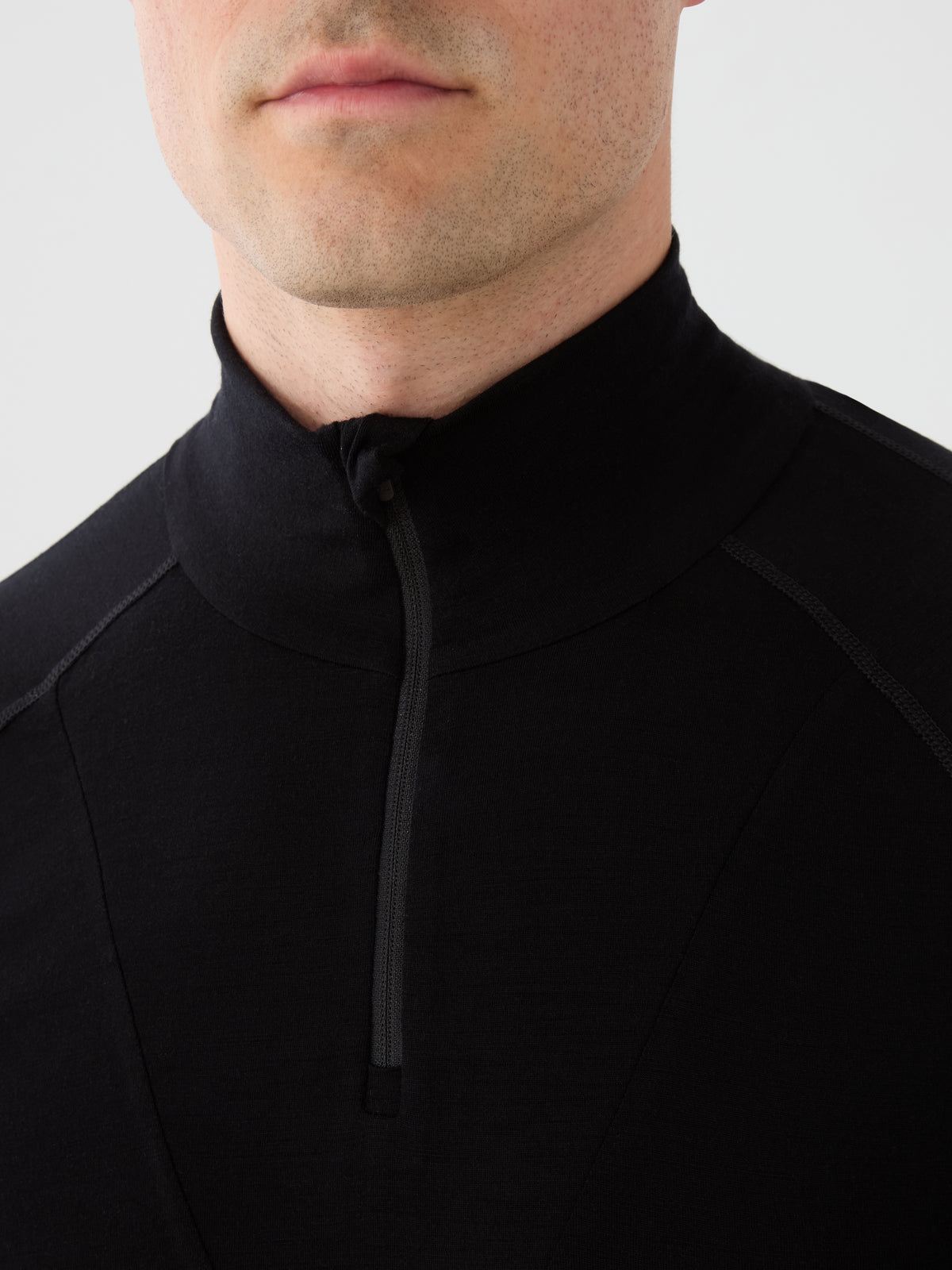 A close-up of a person wearing the We Norwegians Sno Merino Zip Up Men, a black zip-up high-collar athletic shirt designed as a breathable thermal layer, showing only the lower face, neck, and upper chest.
