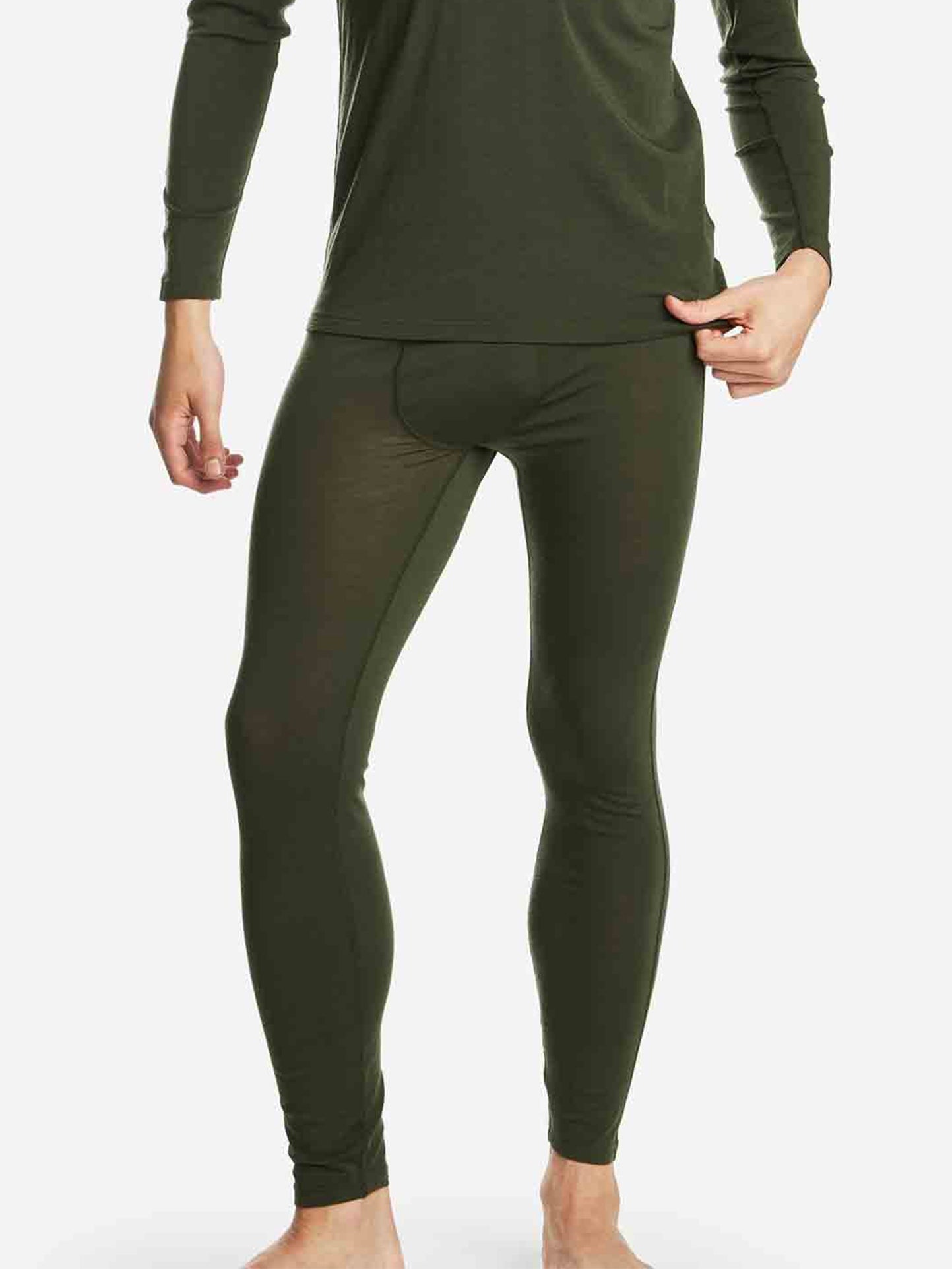 A person wearing We Norwegians Sno Merino Long Johns Bottoms Men in dark green and a matching base-layer long-sleeve top stands barefoot against a plain white background, one hand in their pocket.