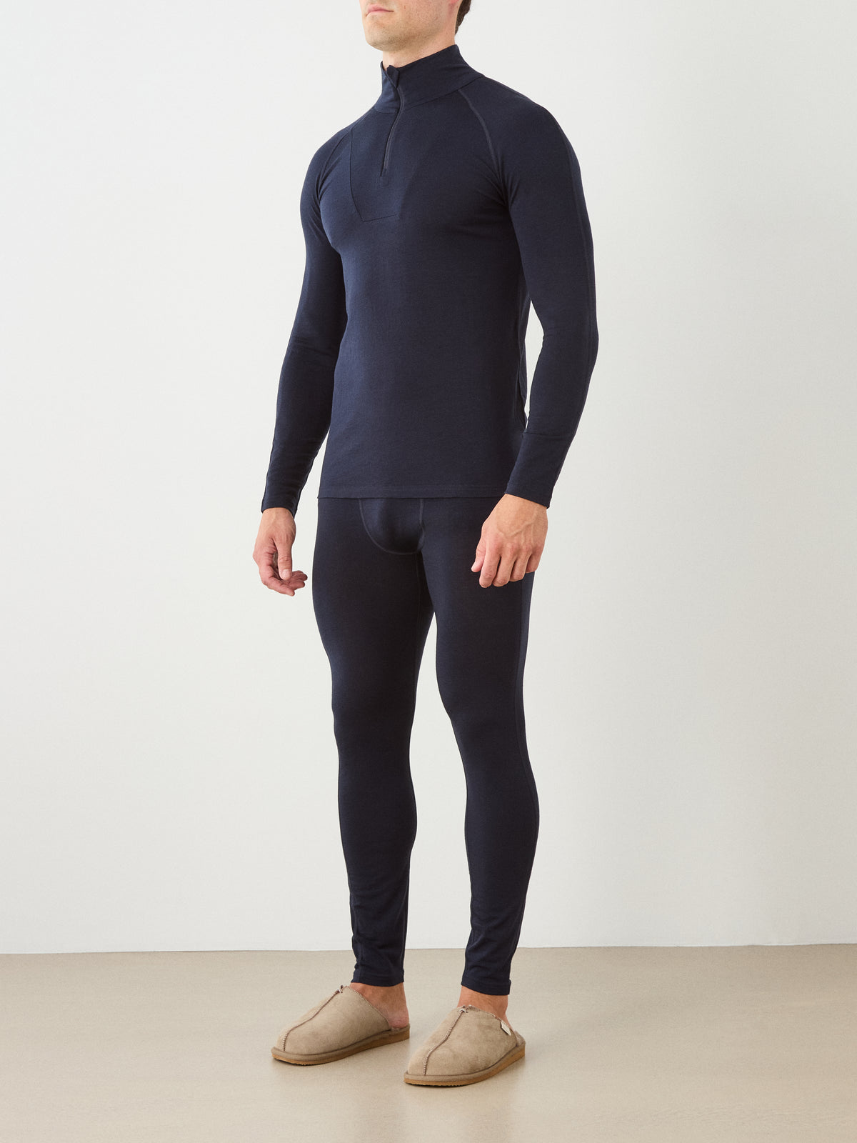 A person stands against a plain indoor background wearing the We Norwegians Sno Merino Zip Up Men—a fitted, long-sleeve navy blue merino wool base layer top—paired with matching leggings and tan slippers.