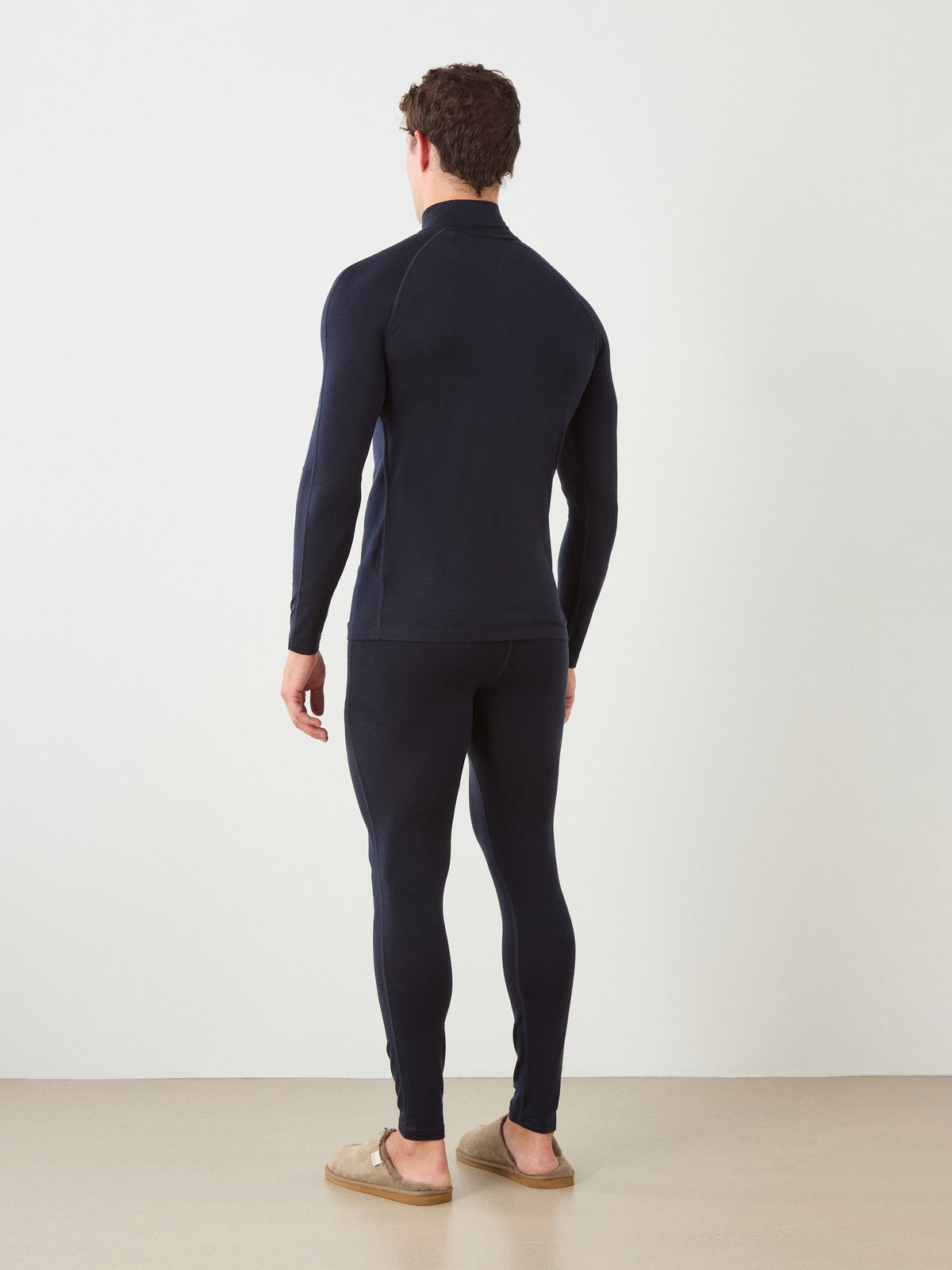 A man stands facing away, wearing the We Norwegians Sno Merino Zip Up Men long-sleeve top and matching leggings in navy with tan slippers. The background is plain and light-colored.