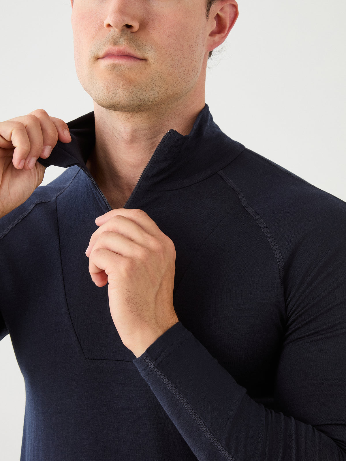 A person is wearing the We Norwegians Sno Merino Zip Up Men, a dark, long-sleeve base layer, and is partially unzipping the collar. Only the lower half of their face and upper torso are visible against a plain light background.