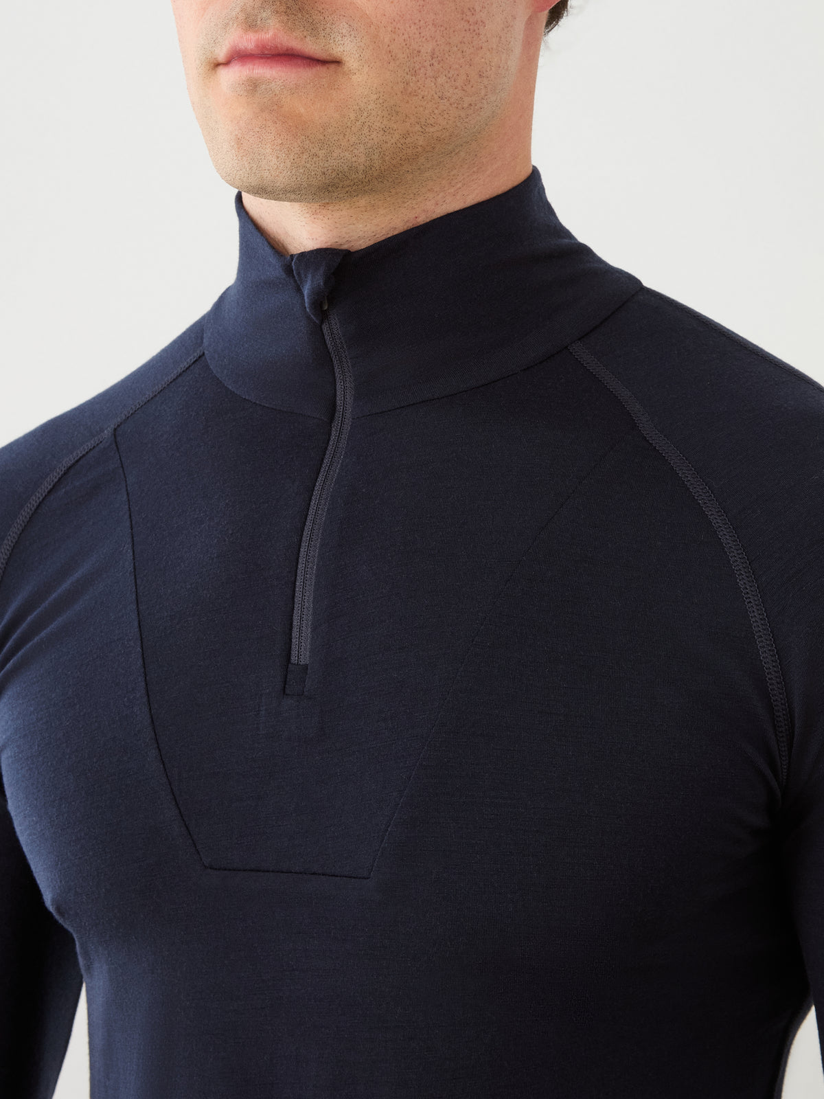 A person wearing the We Norwegians Sno Merino Zip Up Men—a fitted, dark long-sleeve thermal top featuring a high collar and half-zipper—designed for breathability, shown from the chest to just above the mouth.