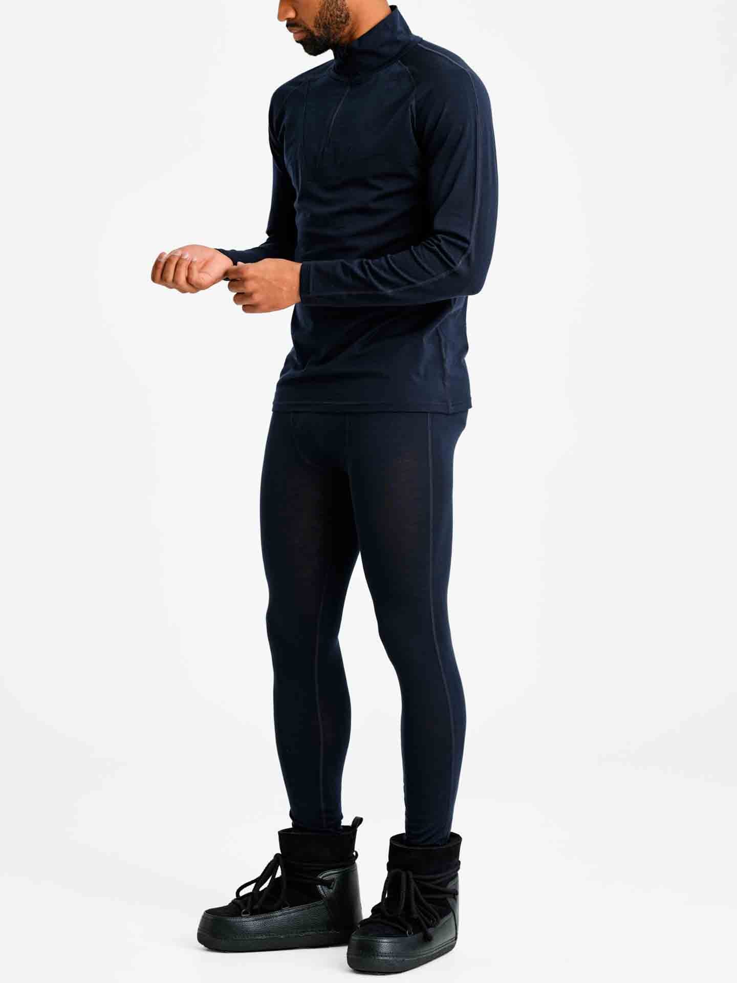 A man dressed in the Sno Merino Zip Up Men from We Norwegians, accompanied by matching navy blue leggings, both crafted from breathable merino wool. He pairs the outfit with black high-top shoes and adjusts a watch on his wrist while standing against a plain white background, exemplifying true Scandinavian luxury base-layers.