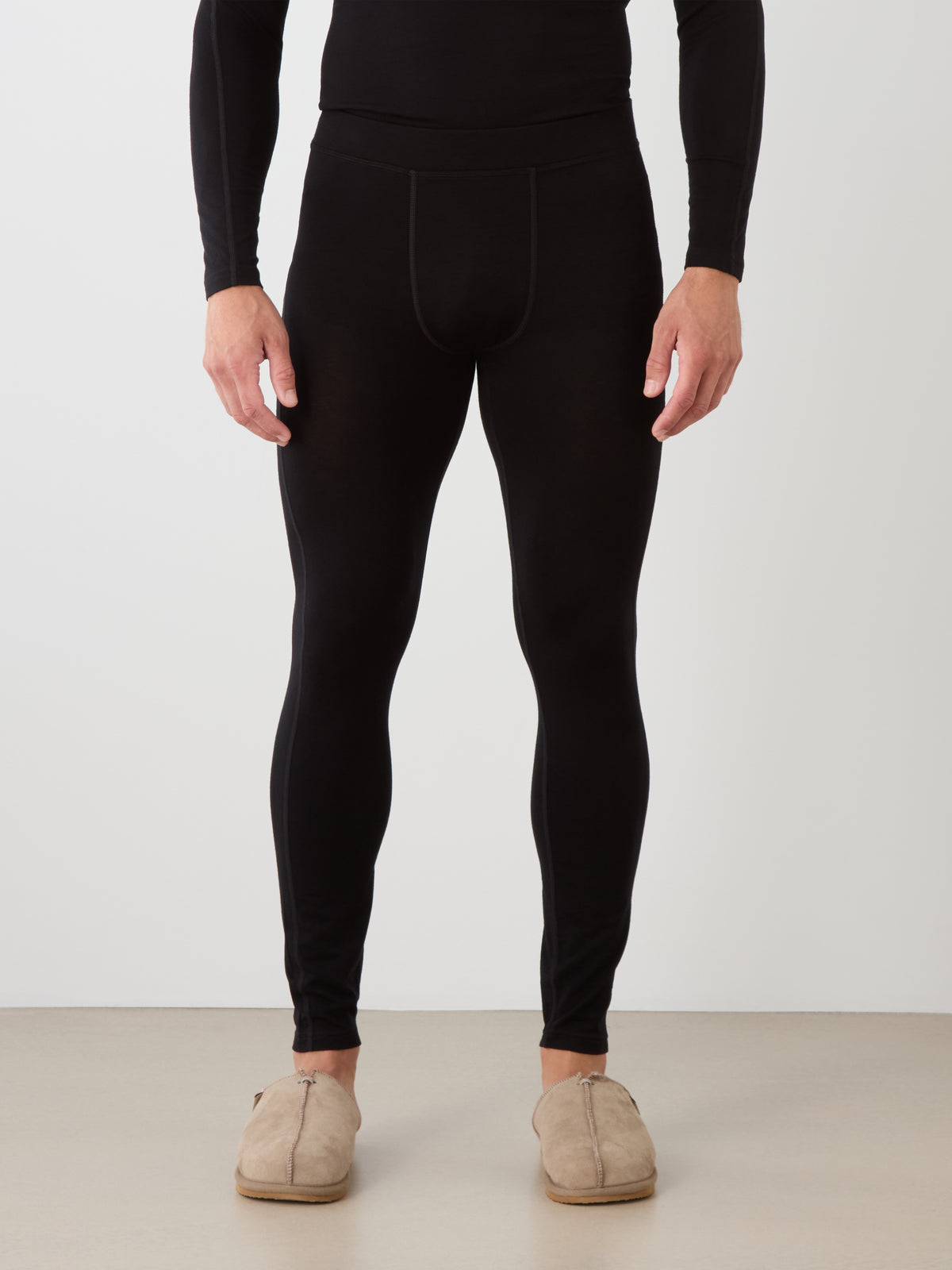 A person wears We Norwegians Sno Merino Long Johns Bottoms Men in black and beige slippers, standing against a plain white wall with their hands at their sides; only the lower body is visible.