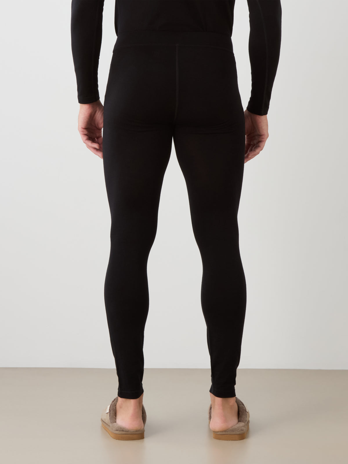 A person stands facing away in black We Norwegians Sno Merino Long Johns Bottoms Men and a breathable long-sleeve base layer, paired with brown slippers, against a plain light background.