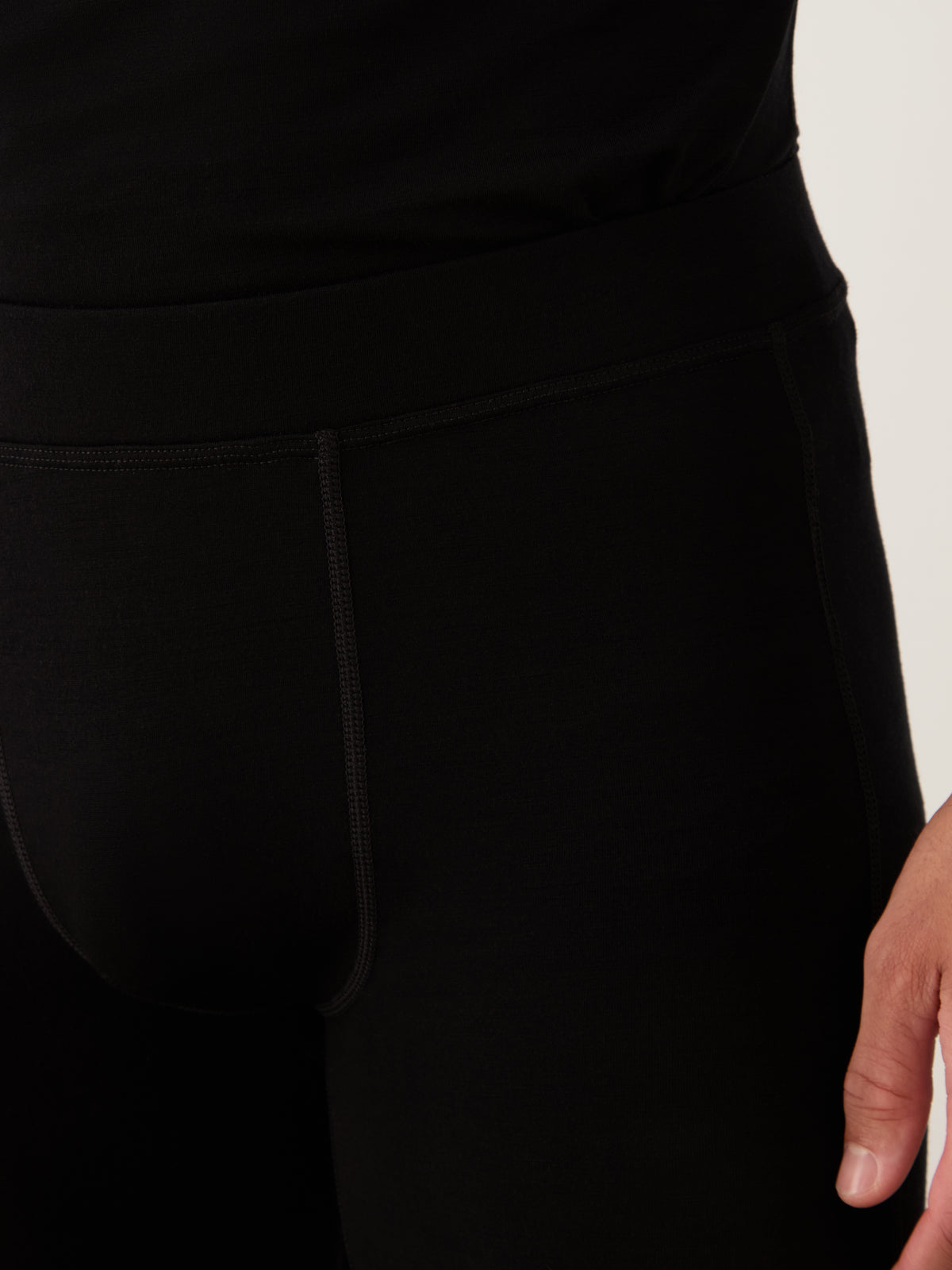 A person wears We Norwegians Sno Merino Long Johns Bottoms Men in black, paired with a matching top. Only the lower torso and one hand are shown against a neutral background.