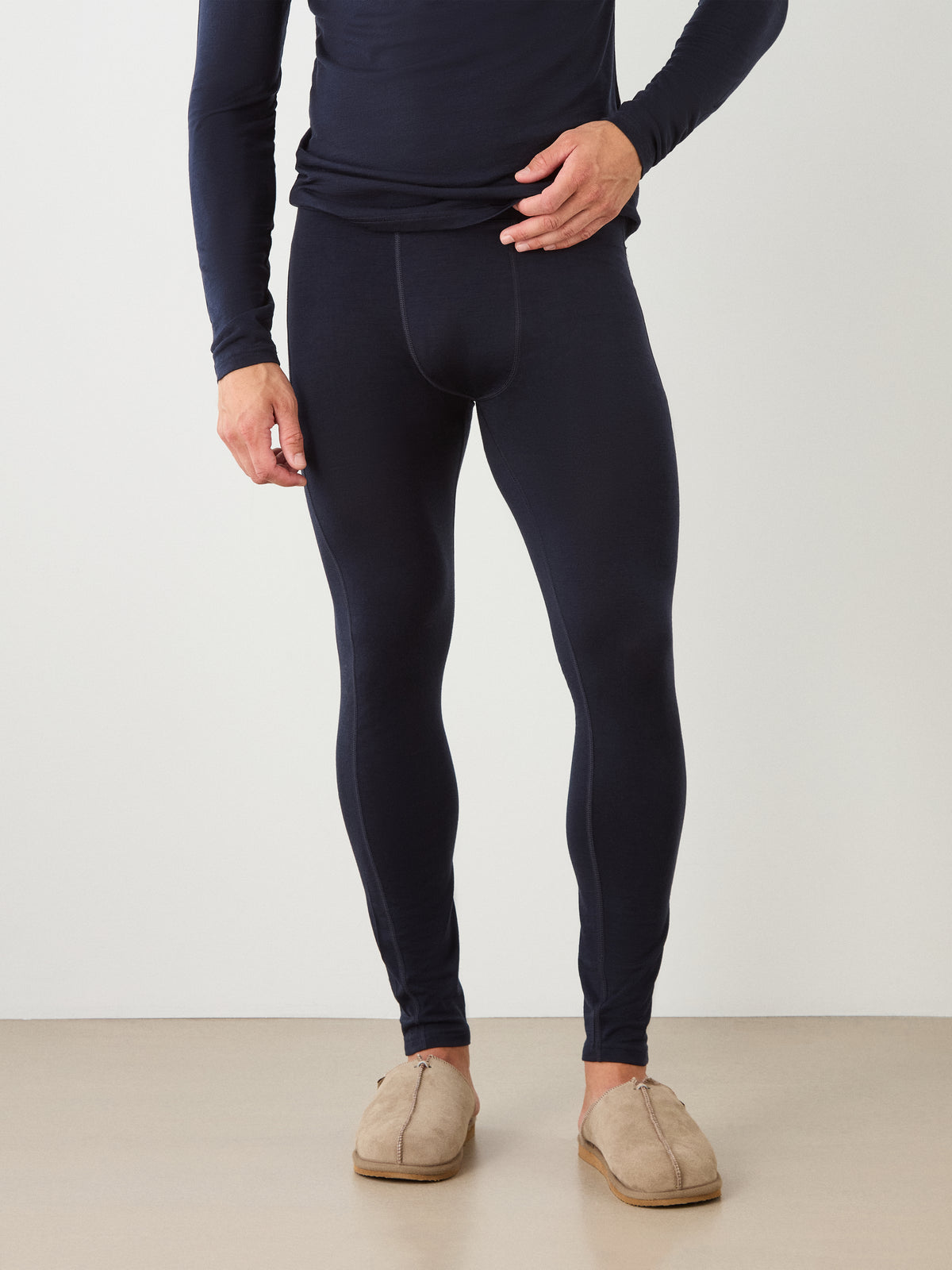 A person wearing We Norwegians Sno Merino Long Johns Bottoms Men in navy blue with a matching long-sleeve top and tan slippers, standing against a plain light-colored wall.
