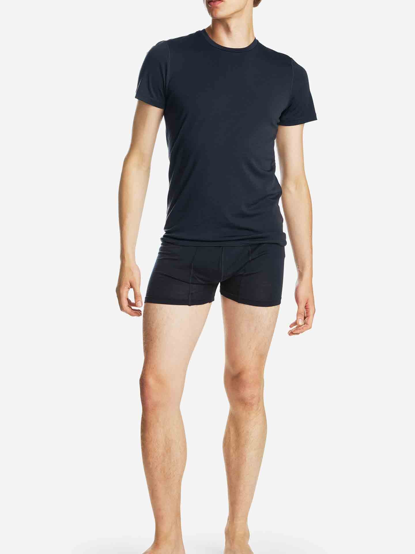 A person wearing the Sno Merino Tee Men by We Norwegians and black boxer briefs, made from Creora-Eco-Soft, stands barefoot against a plain white background. The individual is standing upright, with one arm slightly bent and their head tilted slightly to one side.