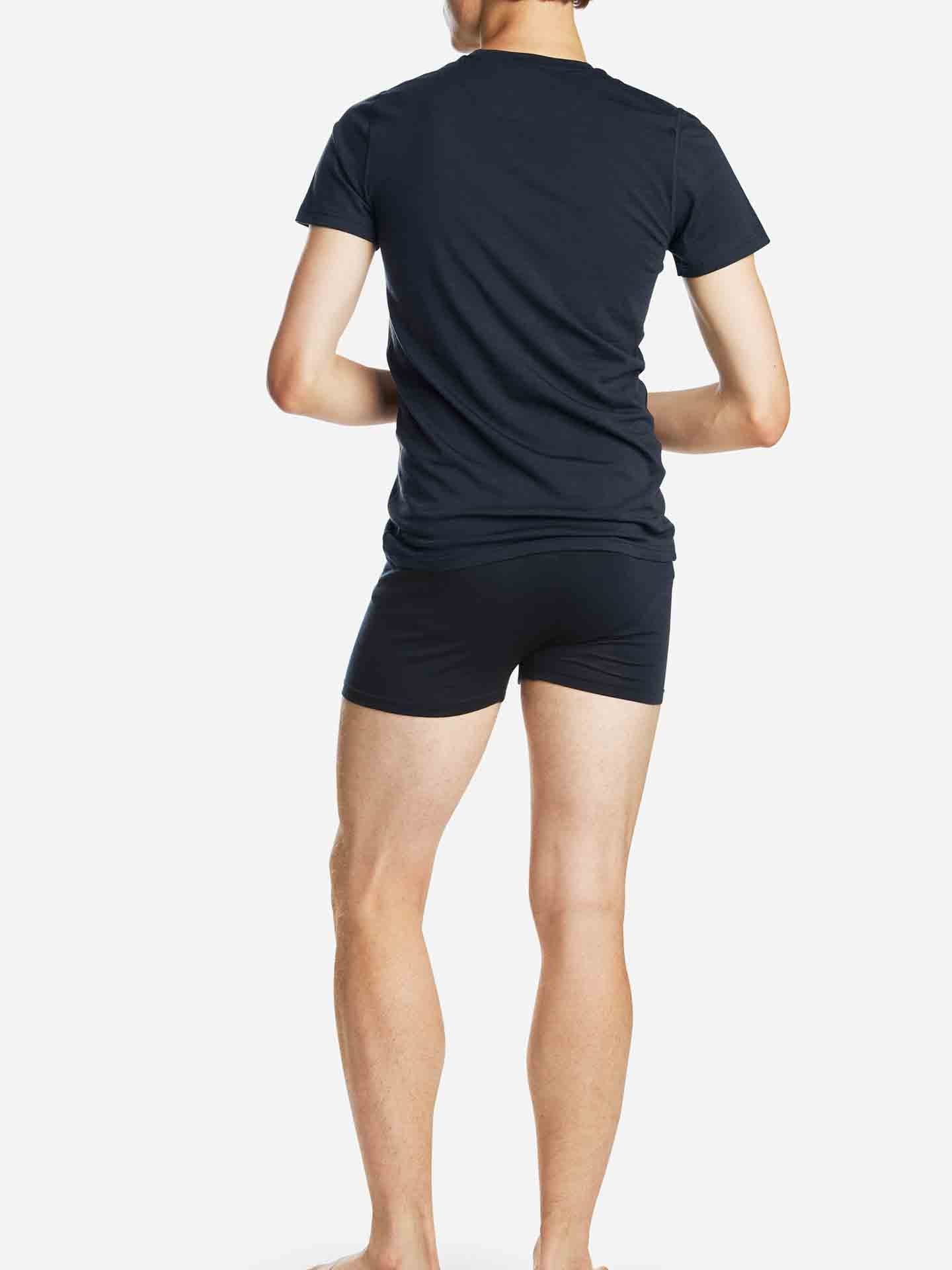 A person is standing barefoot, facing away from the camera, wearing a matching dark-colored "Sno Merino Tee Men" t-shirt and shorts from We Norwegians, made from Creora-Eco-Soft. The background is plain white.