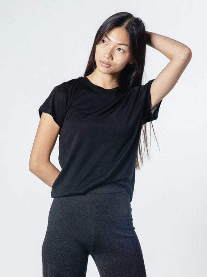 A person with long hair is wearing their favorite We Norwegians Base One Favorite Tee Women and dark pants, standing against a light background. Their right hand is resting on the back of their head while their left arm is relaxed by their side. The expression on their face appears contemplative.