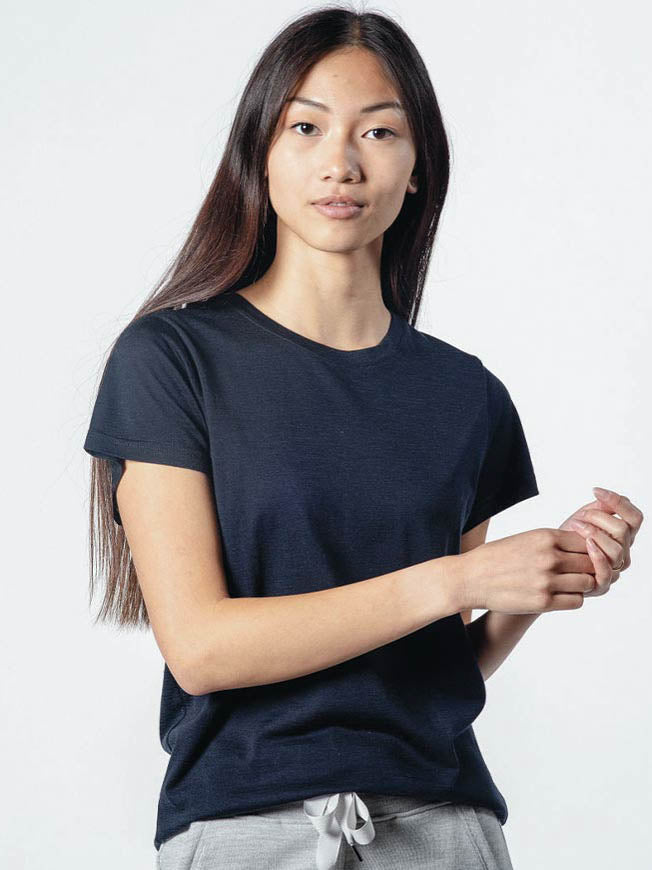 A person with long, straight hair, dressed in their favorite Base One Favorite Tee Women by We Norwegians and light-colored pants, stands against a plain white background. They are looking directly at the camera with a neutral expression, holding their hands gently together in front of them.