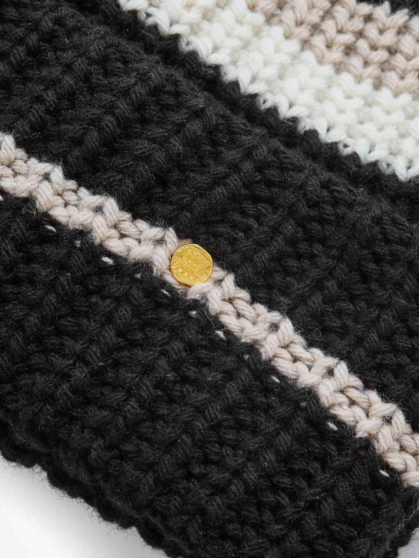 Close-up of the Apres Ski Hat Women by We Norwegians, showcasing a textured pattern in black and beige. The hat features alternating stripes of black, beige, and off-white that highlight its luxurious merino cashmere blend. A small, round, shiny gold emblem or button embellishes the fabric for an elegant touch.