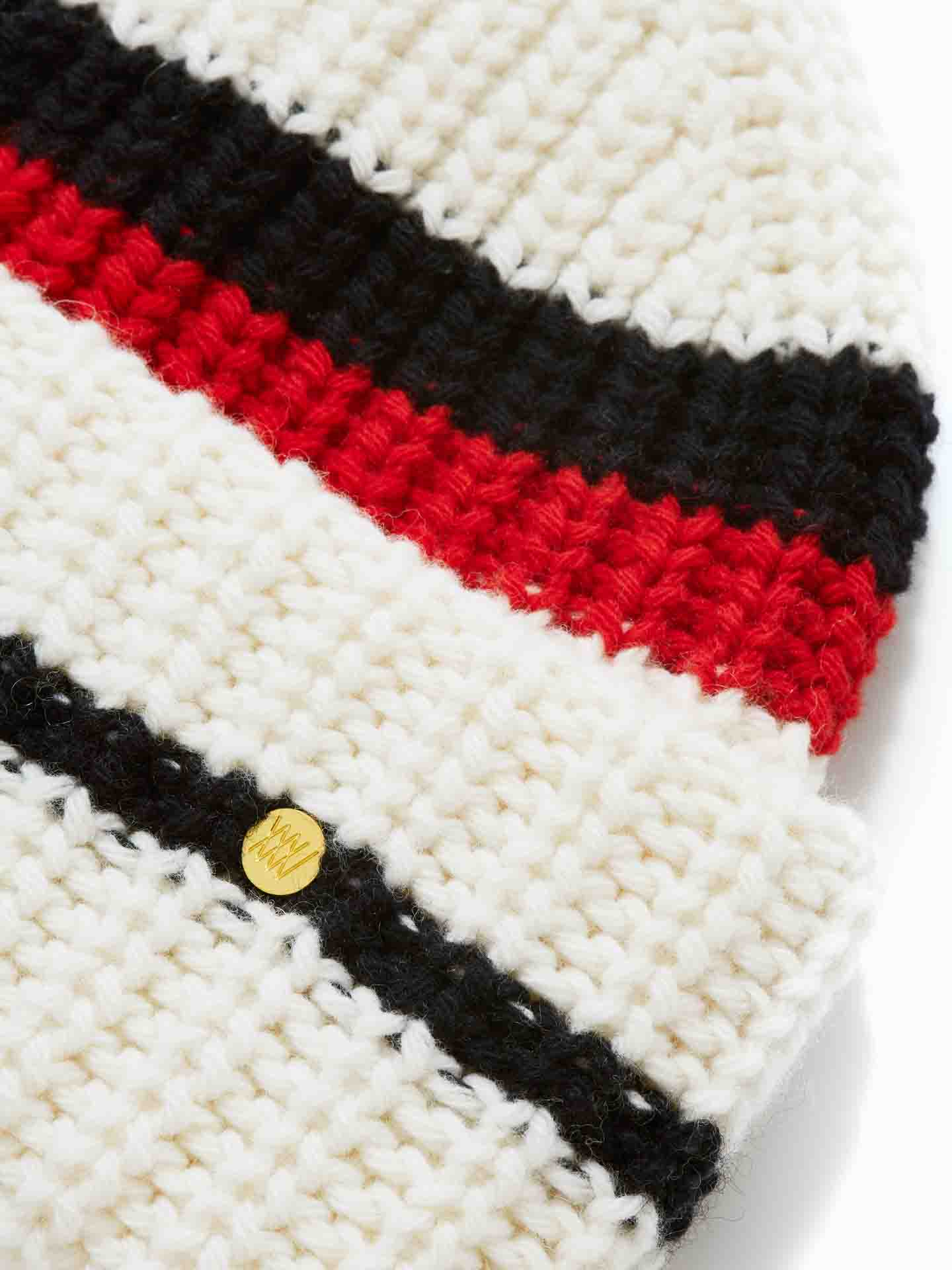 Close-up of an Apres Ski Hat Women by We Norwegians featuring horizontal stripes in black, red, and white. A small circular gold pin with an engraved design is attached to the black stripe. The texture of the chunky knit merino cashmere blend is clearly visible, adding a luxurious touch reminiscent of 70's ski fashion.