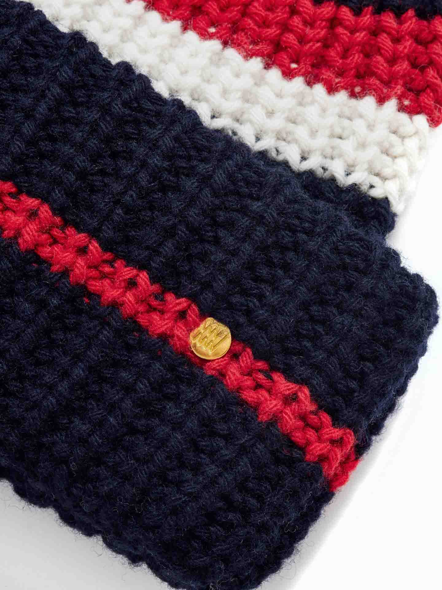 Close-up of a We Norwegians Après Ski Hat Women with red, white, and black yarn stripes. A small round golden tag with a stylized logo is attached to the red section, indicating the brand's mark. The knit texture appears thick and cozy, reminiscent of 70s ski fashion.