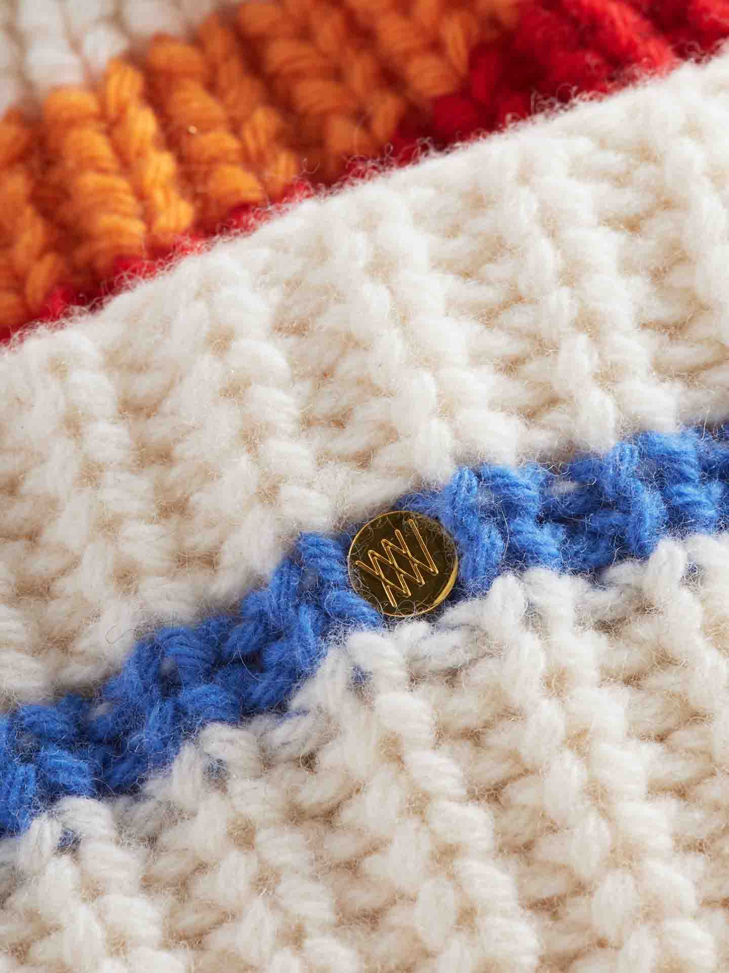 Close-up of the Apres Ski Hat Women by We Norwegians, featuring thick white yarn with blue and orange horizontal stripes. A small, round gold button with a zigzag pattern is attached near the center of the blue stripe. The texture of the knitting, reminiscent of 70's ski fashion, is clearly visible.